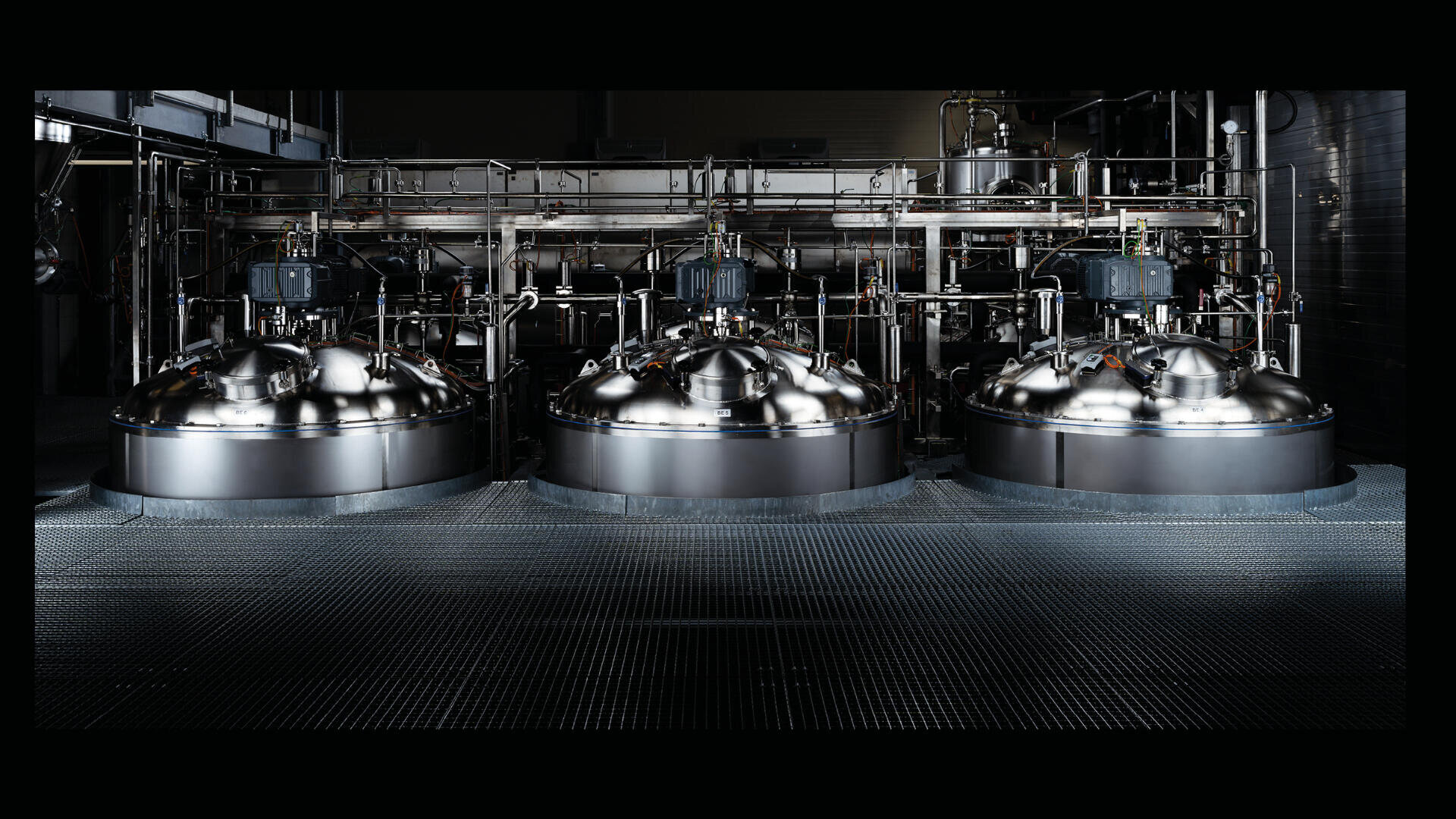 Drei große, glänzende Industrietanks aus Edelstahl mit Rohren, Ventilen und Maschinen in einer schwach beleuchteten Fabrik oder einem Verarbeitungsbetrieb.