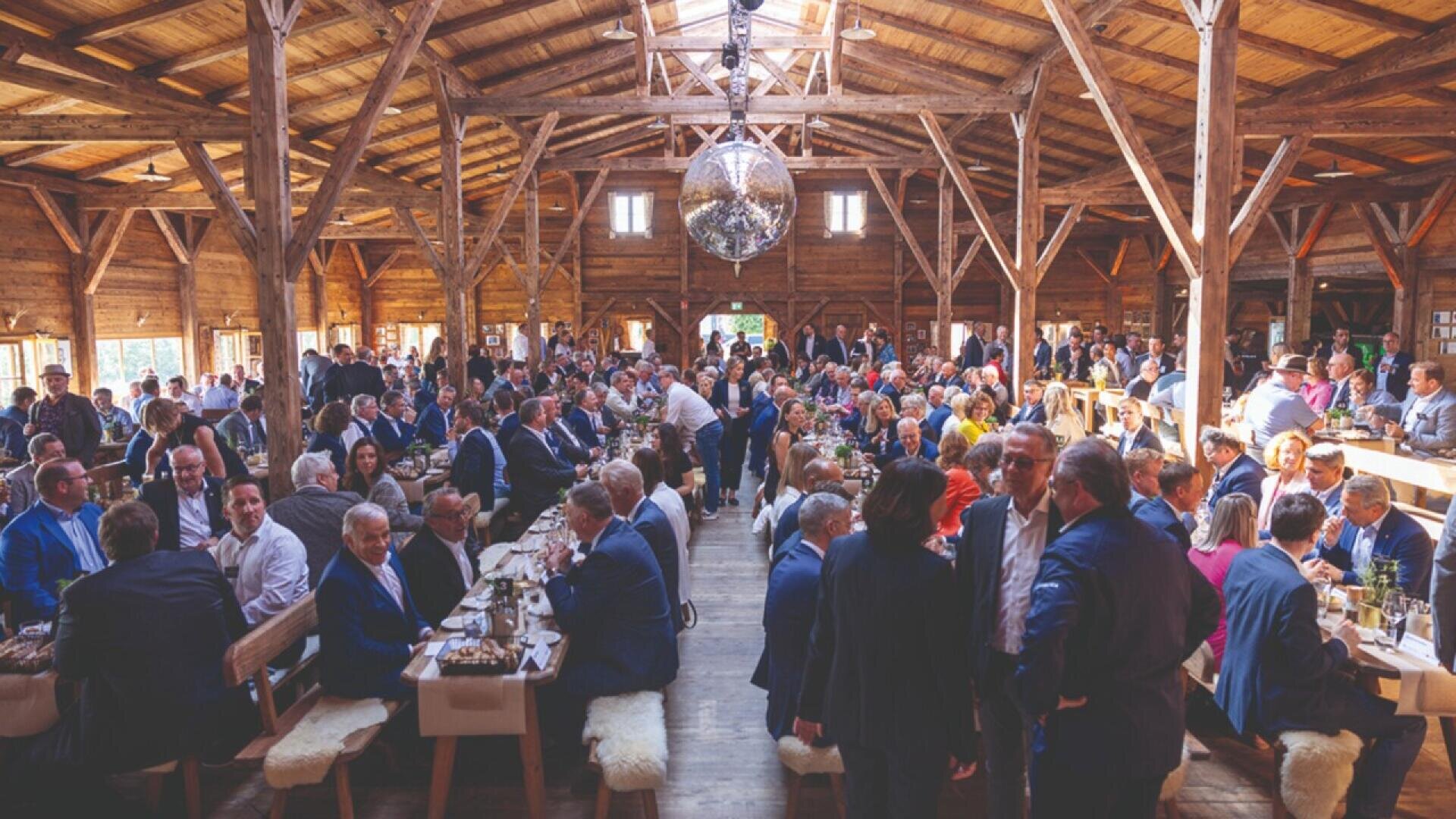 A large group of people dressed in formal attire gather in a rustic wooden hall with exposed beams, sitting at long tables decorated for an event or celebration, under a central disco ball.