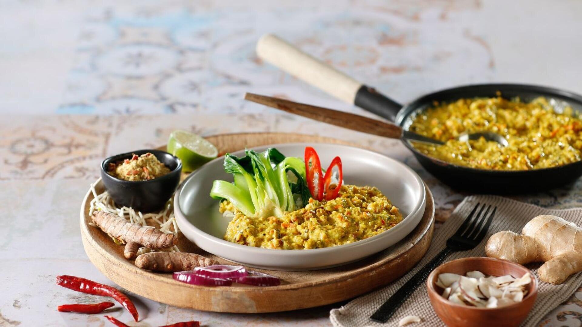 Ein Teller mit gelbem Reis, garniert mit Pak Choi und roten Chilischeiben, wird auf einem Holztablett mit frischem Kurkuma, Ingwer, roten Zwiebeln und Chili serviert, neben kleinen Schüsseln mit Gewürzen und einem Topf Reis im Hintergrund.