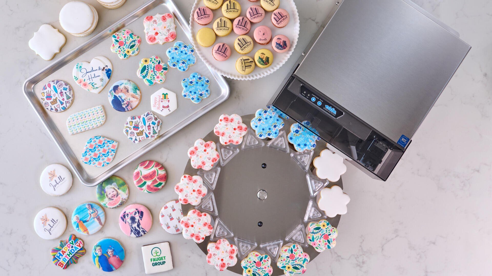 Colorful cookies and macarons decorated with printed images, text, and patterns are arranged on trays and a turntable next to a compact silver food printer on a white countertop.