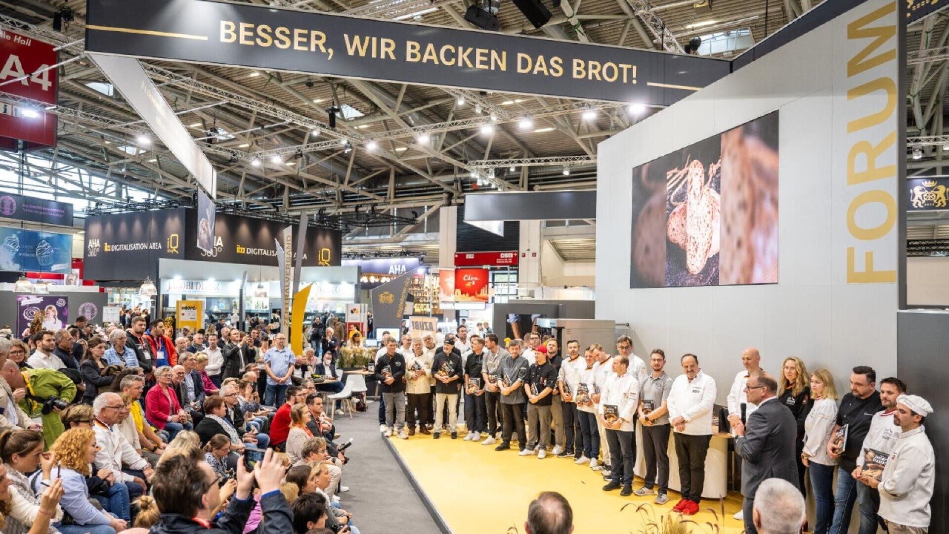 A large audience watches a group of people standing on stage at a trade show or conference. Above them is a banner that reads “BESSER, WIR BACKEN DAS BROT!” and a large screen shows an image of bread.