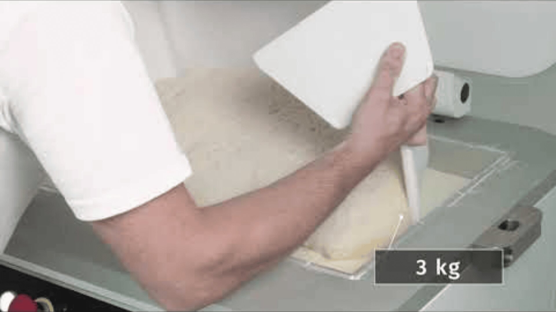 A person in a white shirt cuts dough on a flat surface with a plastic dough scraper. A label in the picture says 3 kg.