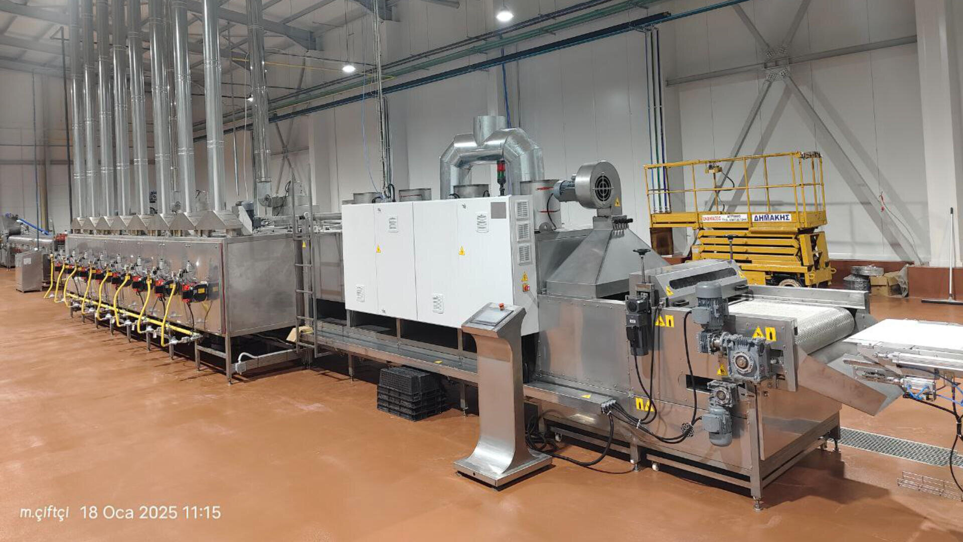 A large, industrial food processing machine with multiple connected stainless-steel units stands in a bright factory room. Nearby, there is a yellow scissor lift. The floor is clean and shiny brown.
