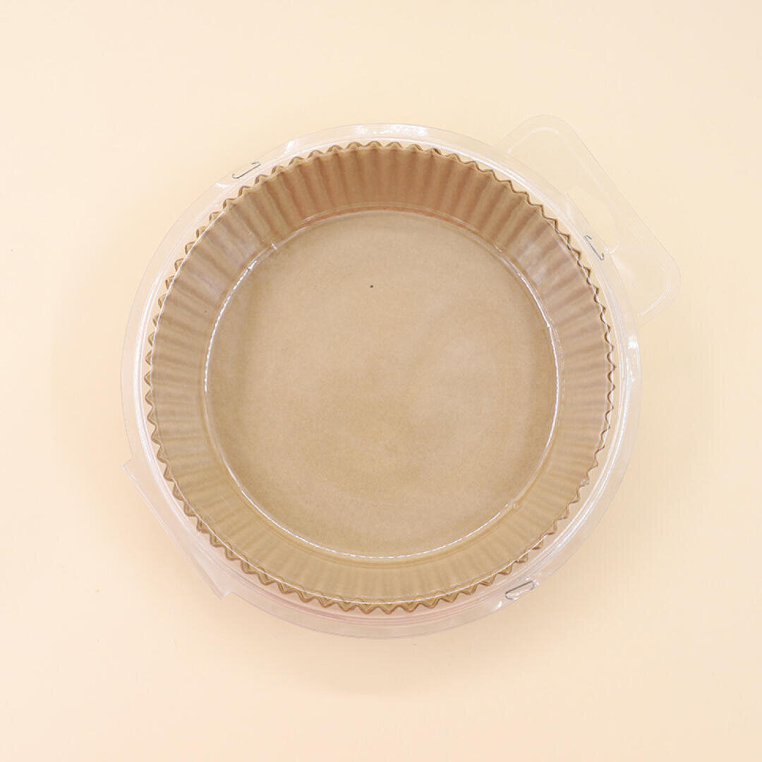 A round, empty, brown paper baking mold with fluted edges, sits inside a clear plastic container on a light beige background.