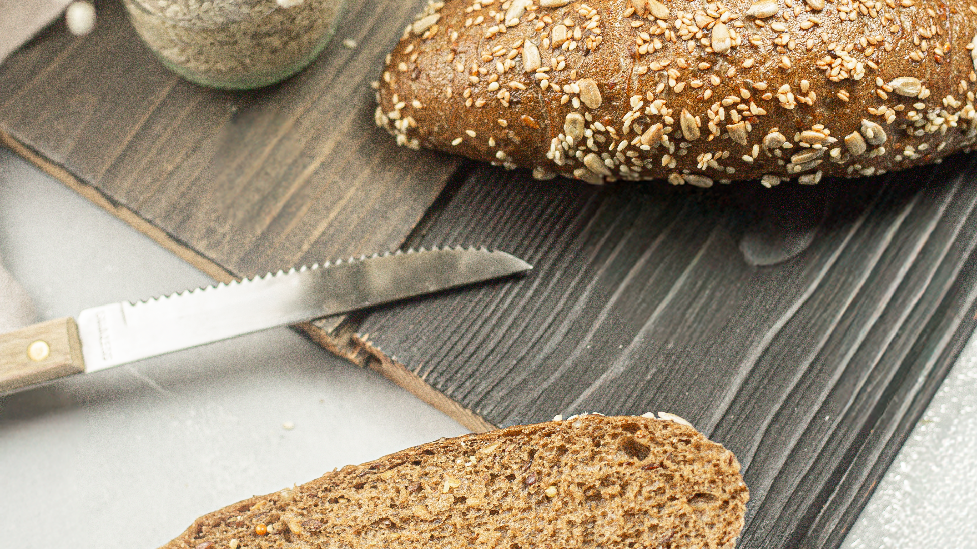 A loaf of seeded whole grain bread sits on a wooden board next to a bread knife and a glass jar of seeds; a sliced piece of bread is visible in the foreground.