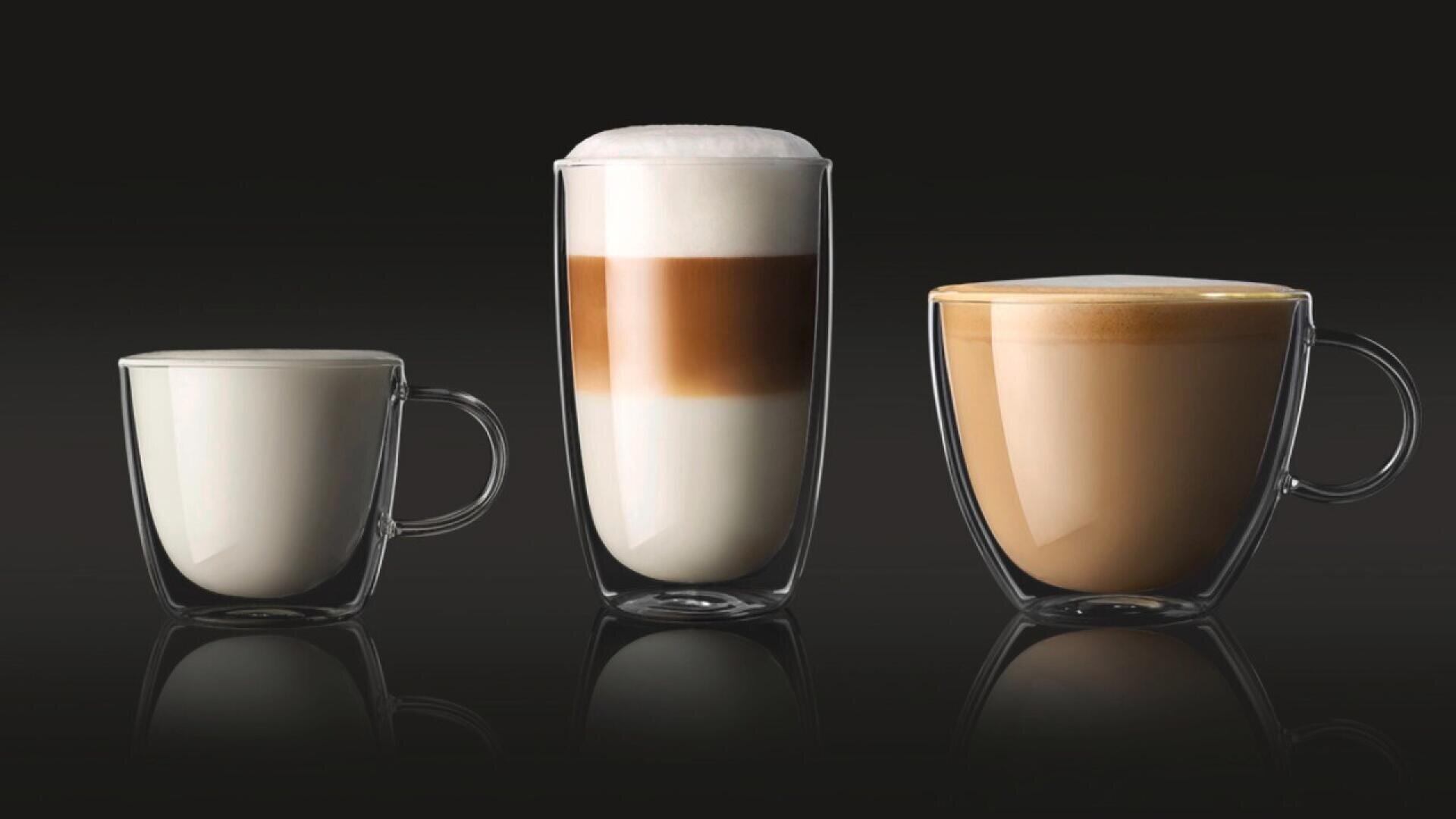 Three clear glass cups with handles are lined up, each containing a different coffee drink: a cup of milk, a layered latte macchiato, and a creamy cappuccino, all set against a dark background.