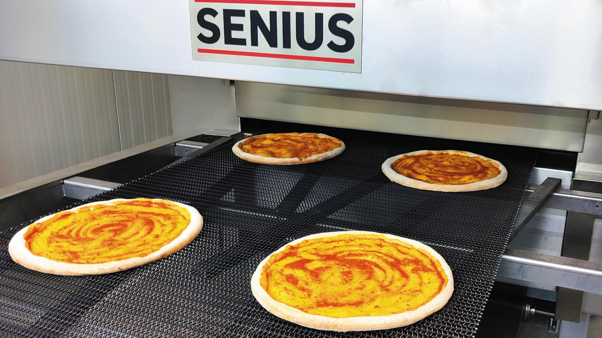 Four uncooked pizza bases with tomato sauce move along a conveyor belt inside an industrial pizza-making machine labeled SENIUS.