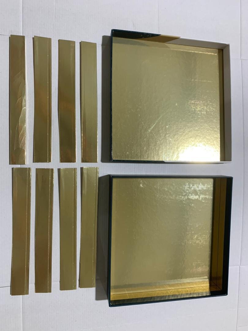 Two open gold-colored rectangular boxes with shiny interiors are placed on a white surface. To the left of the boxes, eight long, narrow gold strips are arranged in two vertical rows.
