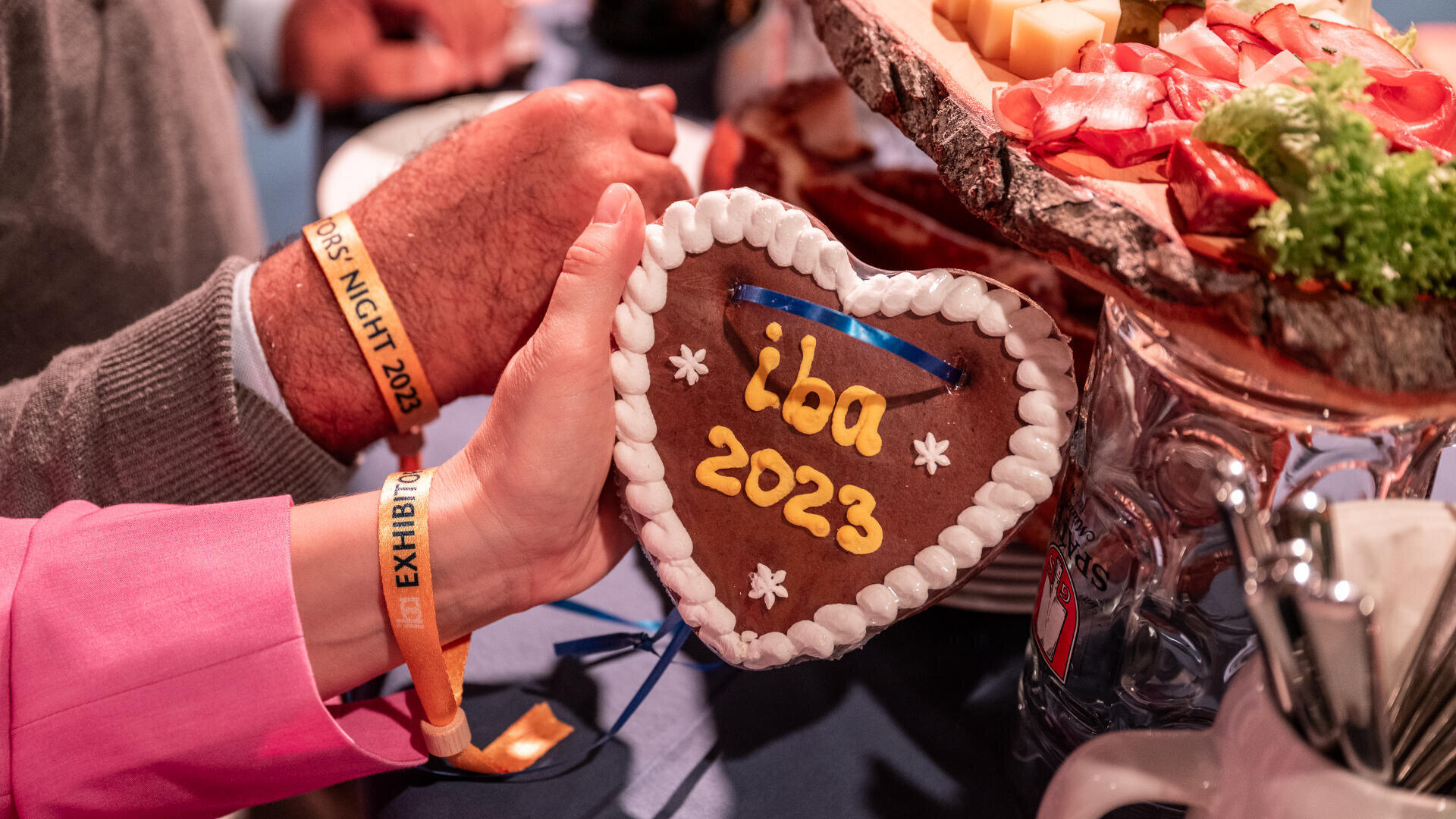 Eine Person hält einen herzförmigen Lebkuchen mit der Aufschrift iba 2023 in gelbem Zuckerguss in der Hand, umgeben von Servierplatten mit Käse und Fleisch. Die Person trägt ein Armband mit der Aufschrift EXHIBITOR.
