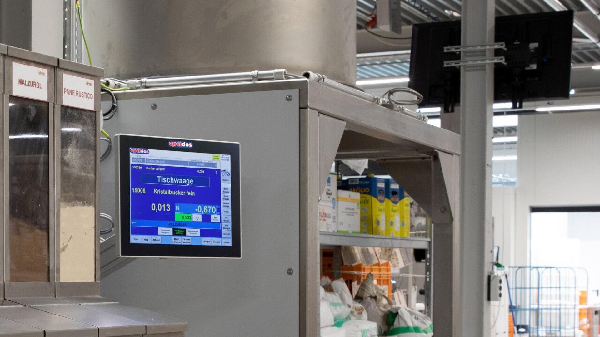 A digital weighing scale with a display screen showing measurements is mounted on a metal structure in an industrial setting, with shelves of supplies and large equipment visible in the background.