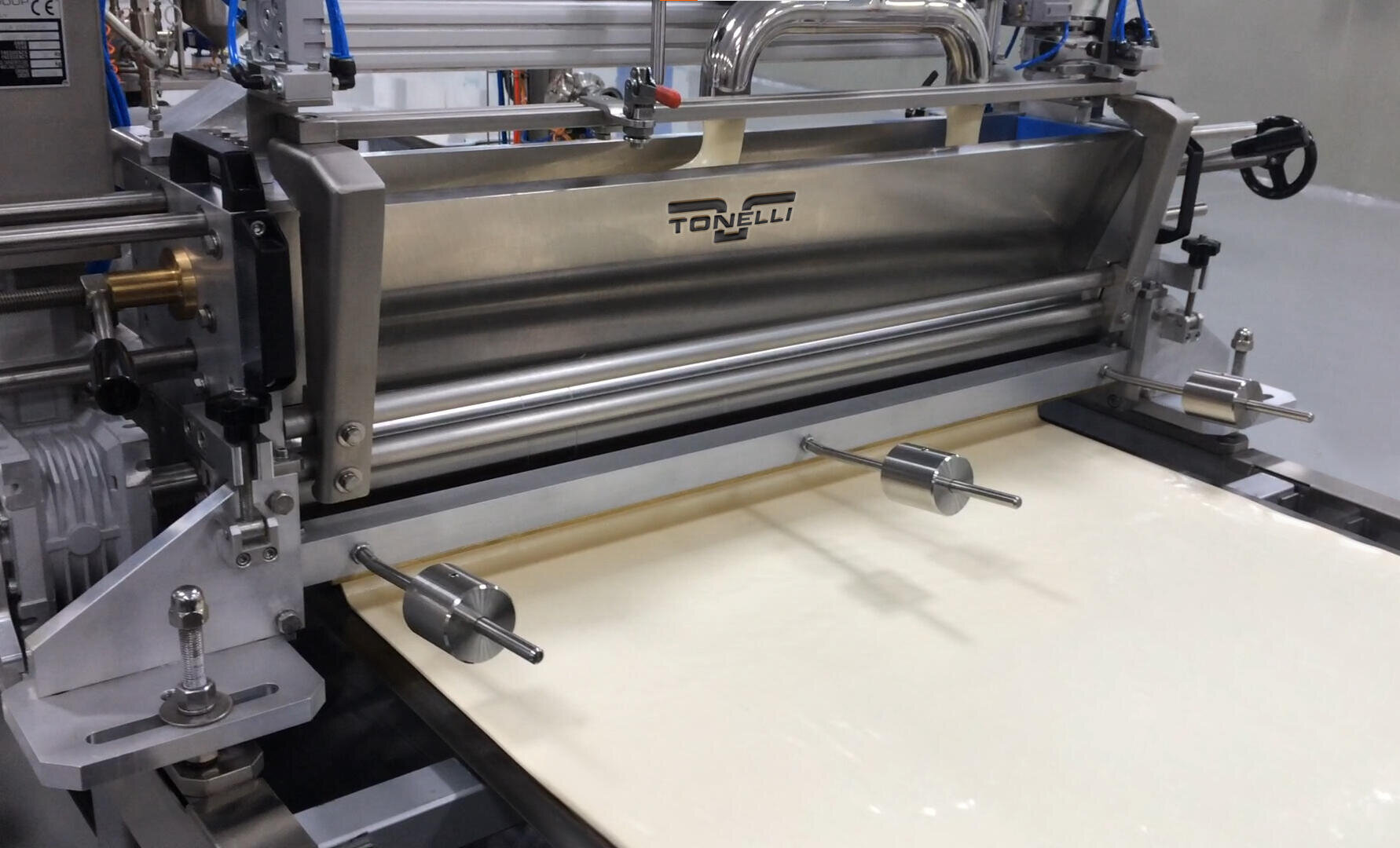 A stainless steel Tonelli industrial machine processes a sheet of dough or batter, rolling it out smoothly on a conveyor in a clean, modern food production facility.