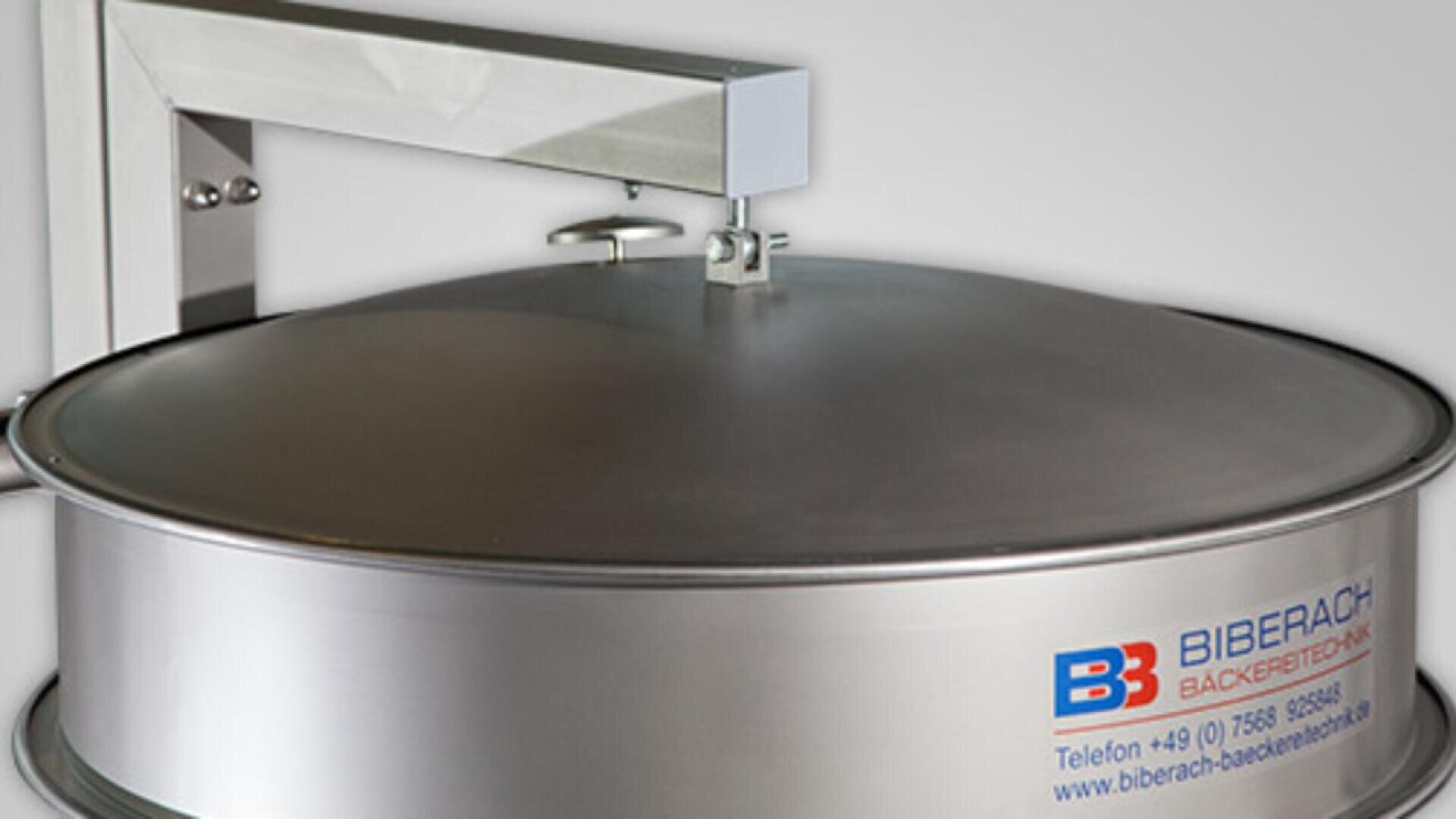 A large stainless steel industrial baking machine with a closed circular lid, labeled Biberach Bäckereitechnik, and features a mounted arm above the lid. Contact information is printed on the front.
