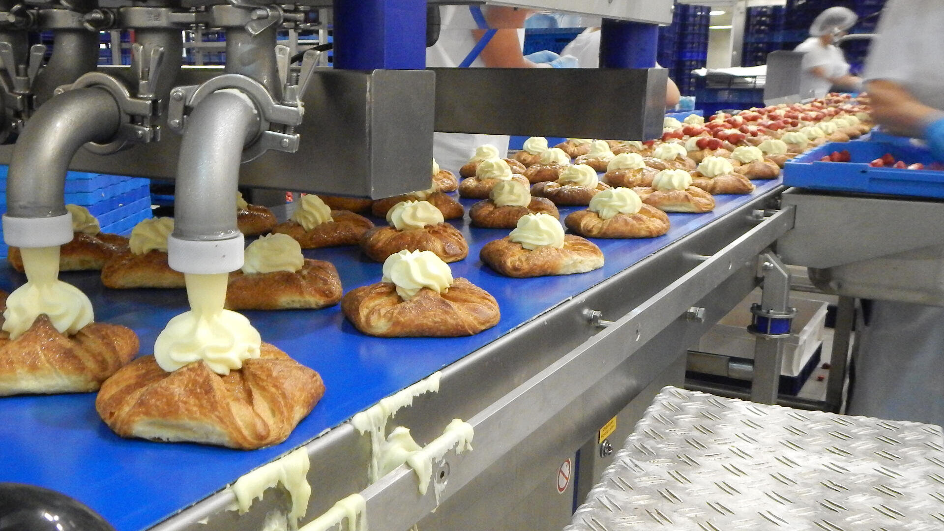 Gebäck läuft über ein blaues Förderband, während eine Maschine Sahne auf das Gebäck aufträgt; im Hintergrund einer Großbäckerei arbeiten Arbeiter in weißen Uniformen mit Backwaren.