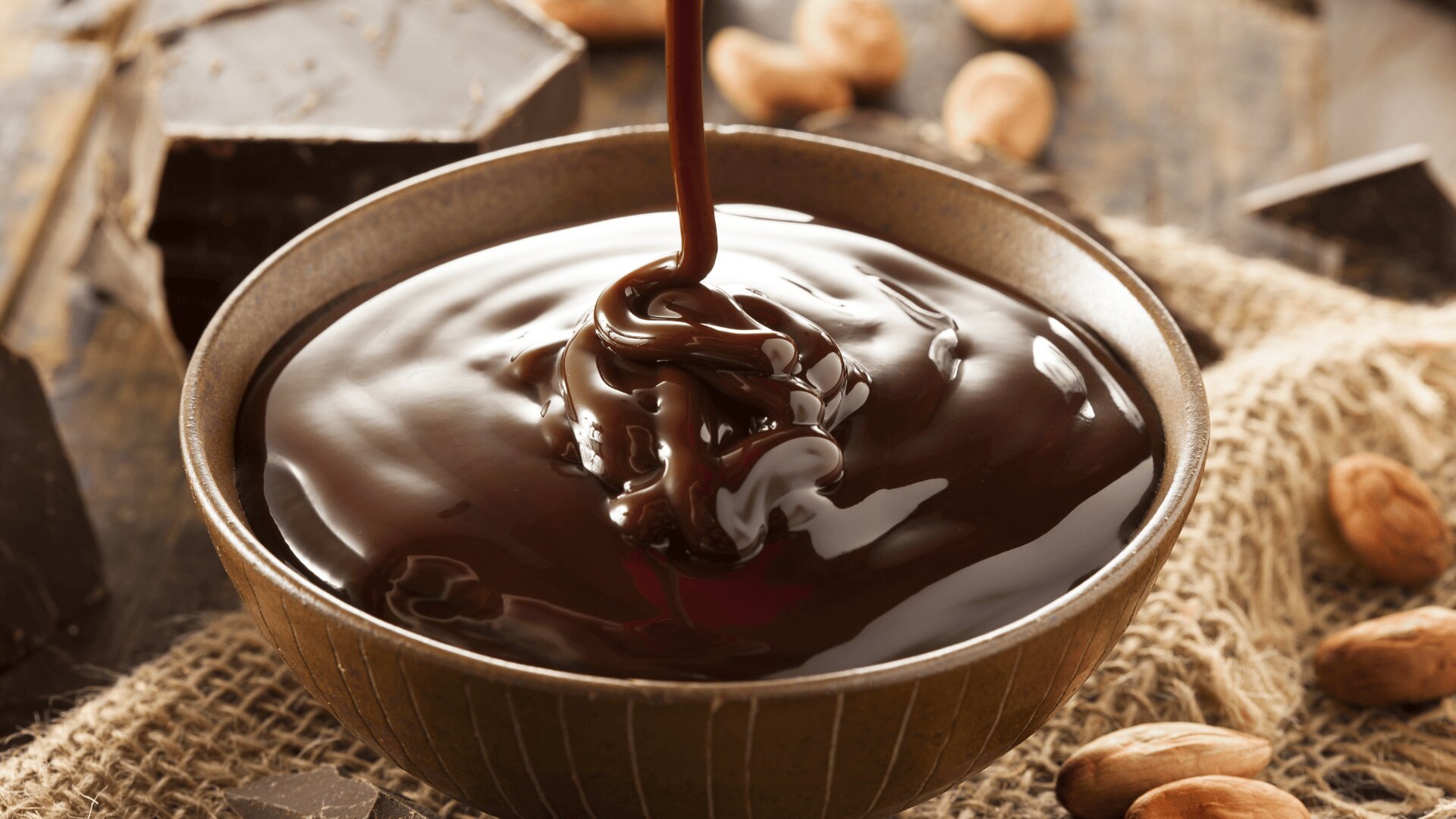 A bowl filled with smooth, melted chocolate being poured in, surrounded by cocoa beans, chocolate pieces, and a burlap cloth.
