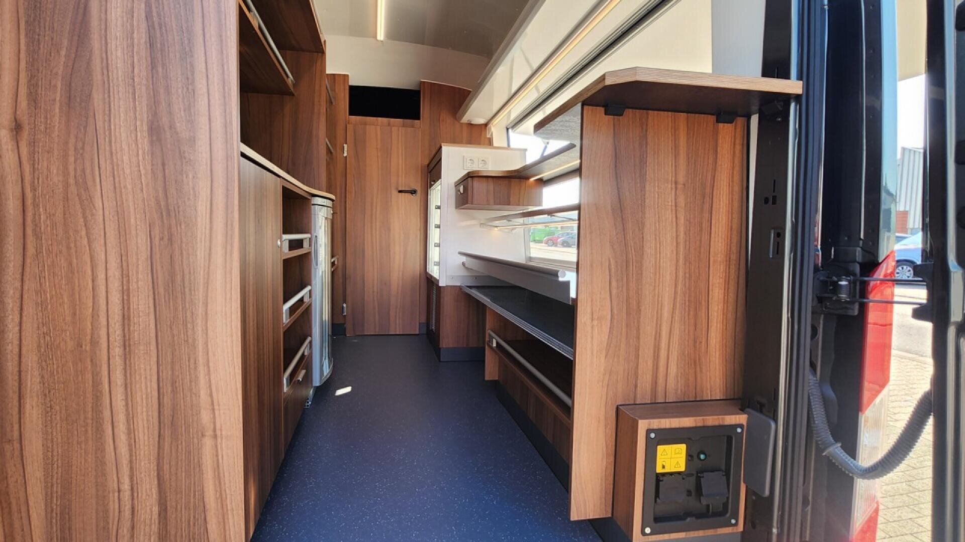 Interior of a mobile workspace or food truck, featuring wood paneling, shelves, drawers, counters, and cabinets along both sides, with natural light entering from open windows on the right.