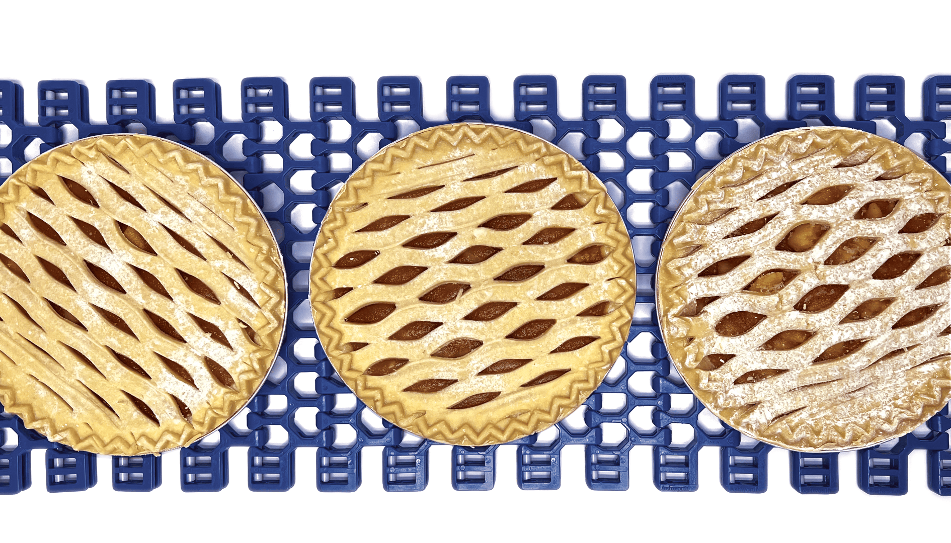 Three lattice-topped pies sit on a blue conveyor belt, viewed from above. The pies have a golden, crisscrossed pastry pattern and are aligned in a row against a white background.