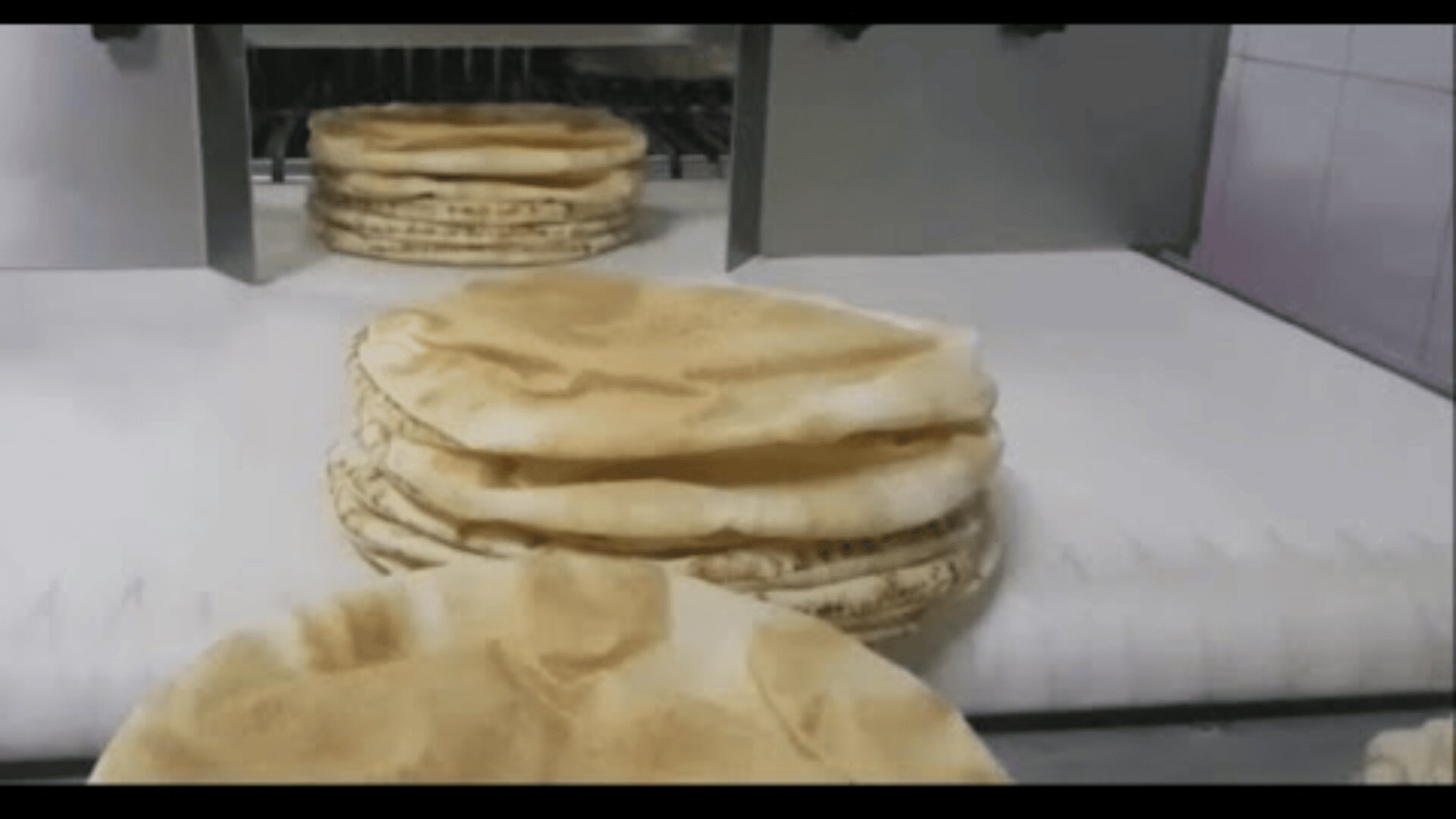 Stacks of round pita bread are moving on a conveyor belt inside a bakery or bread production facility. The bread is light brown and fluffy, freshly baked and ready for packaging.
