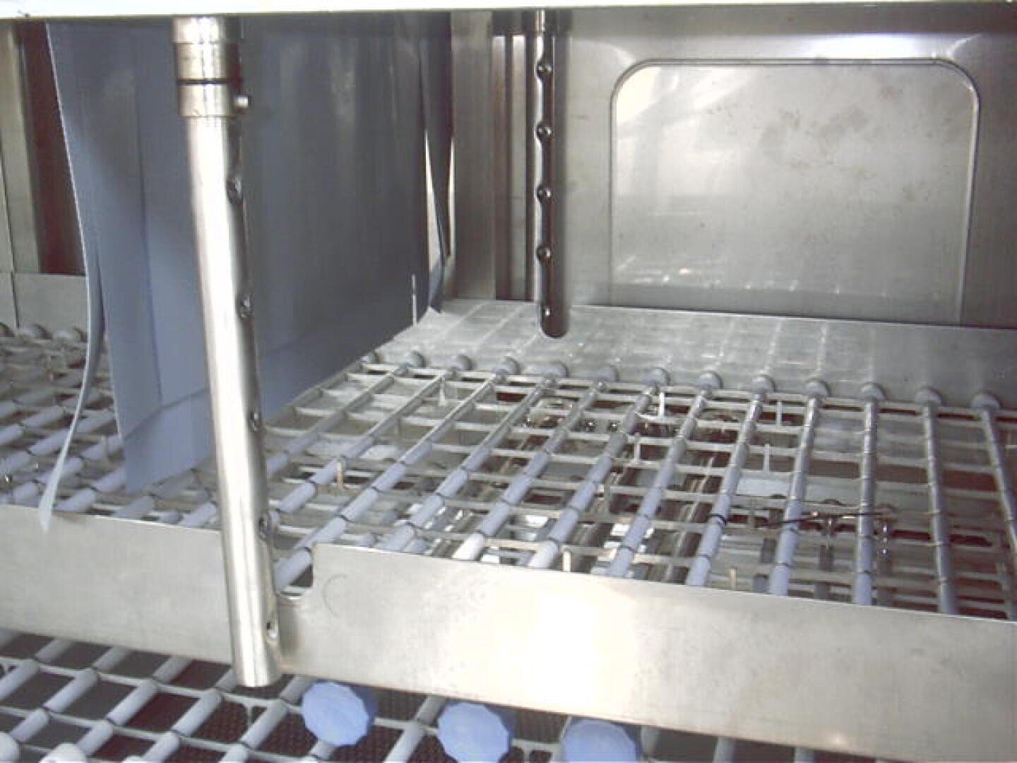 Close-up of an empty dishwasher rack with metal grids, blue plastic components, and vertical spray arms inside a stainless steel dishwasher interior.