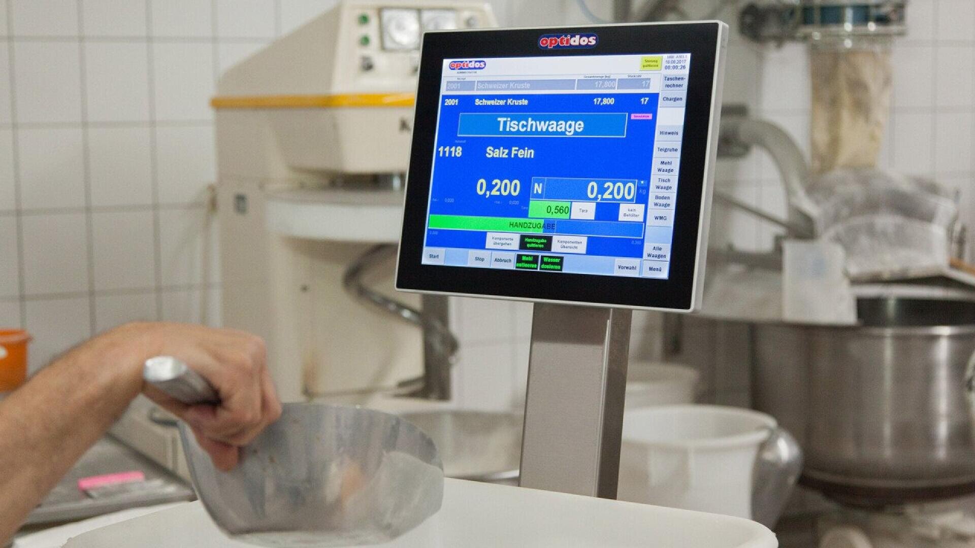 A close-up of an electronic scale display in a kitchen, showing weight measurements in kilograms, with a person scooping fine salt into a container below the scale. Various kitchen equipment is visible in the background.