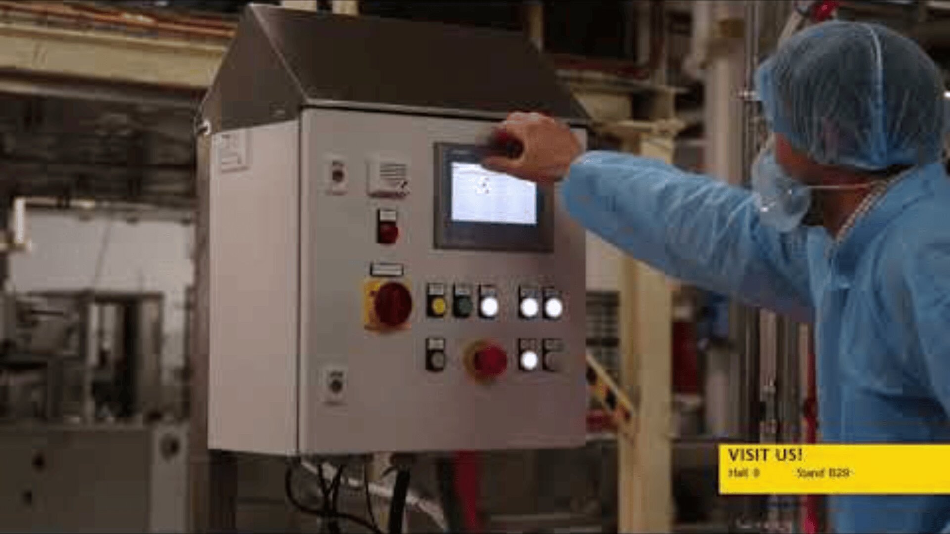 A person in protective clothing and a hairnet is operating an industrial control panel with various buttons and a screen inside a factory setting. A yellow banner in the corner reads, VISIT US! Hall 8 Stand B29.