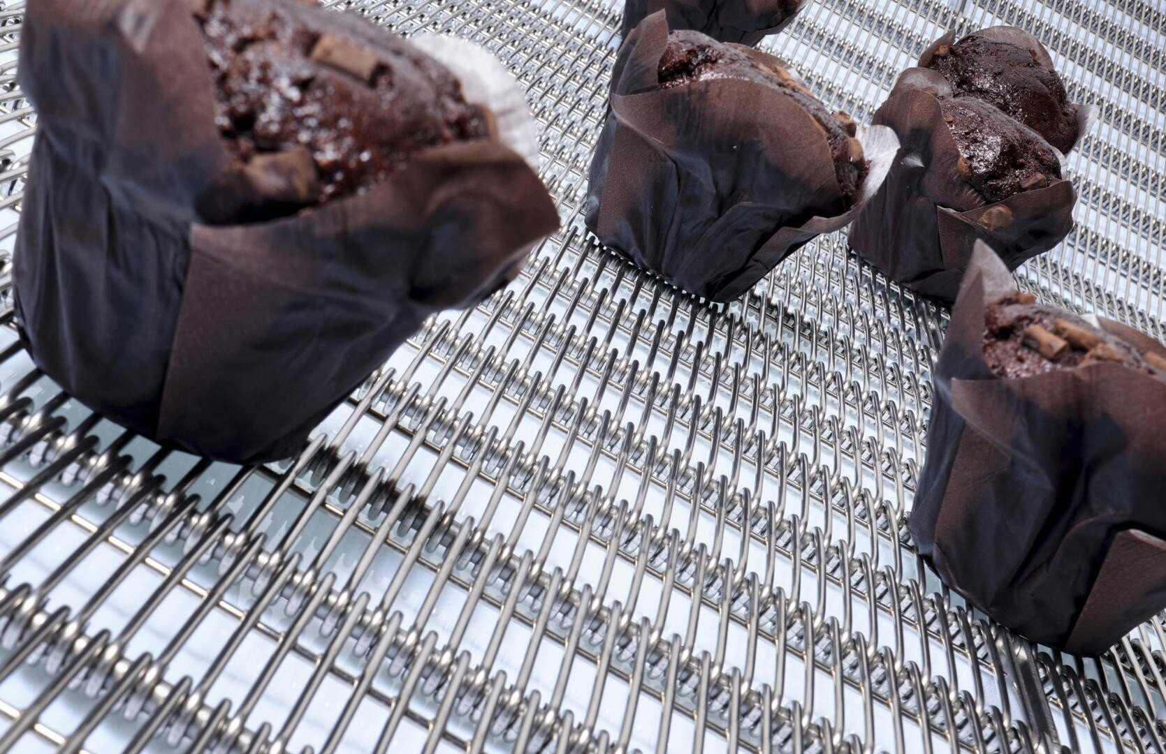 Chocolate muffins in dark paper wrappers are cooling on a metal wire rack, with a close-up and angled perspective.