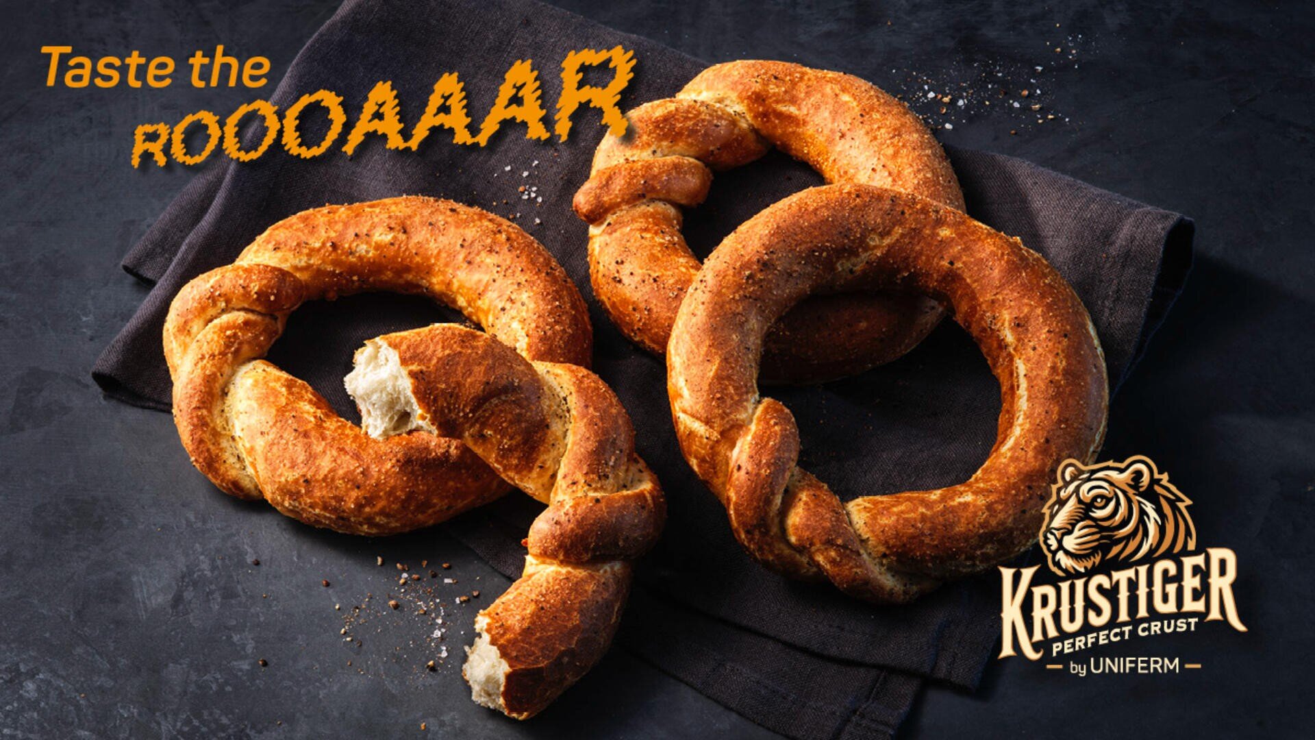 Drei gedrehte Brotringe mit einer goldbraunen, knusprigen Kruste liegen auf einer dunklen Serviette. Der Text lautet: „Taste the ROOOAAAR.“ Das Krustiger-Logo mit einem Tiger ist in der unteren rechten Ecke zu sehen.