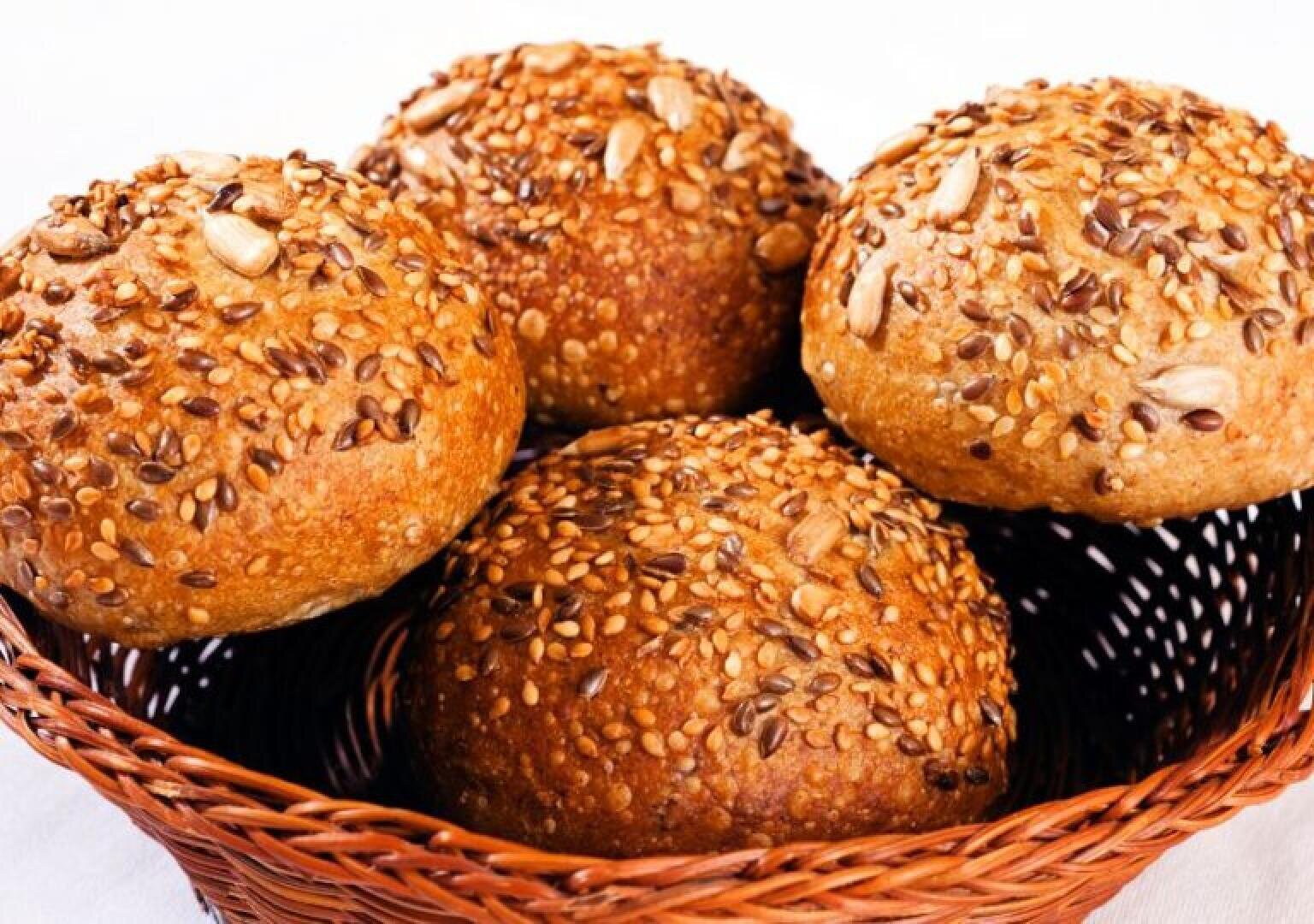 A wicker basket filled with four round, whole grain bread rolls topped with a mix of seeds, including sunflower and sesame seeds, on a white background.