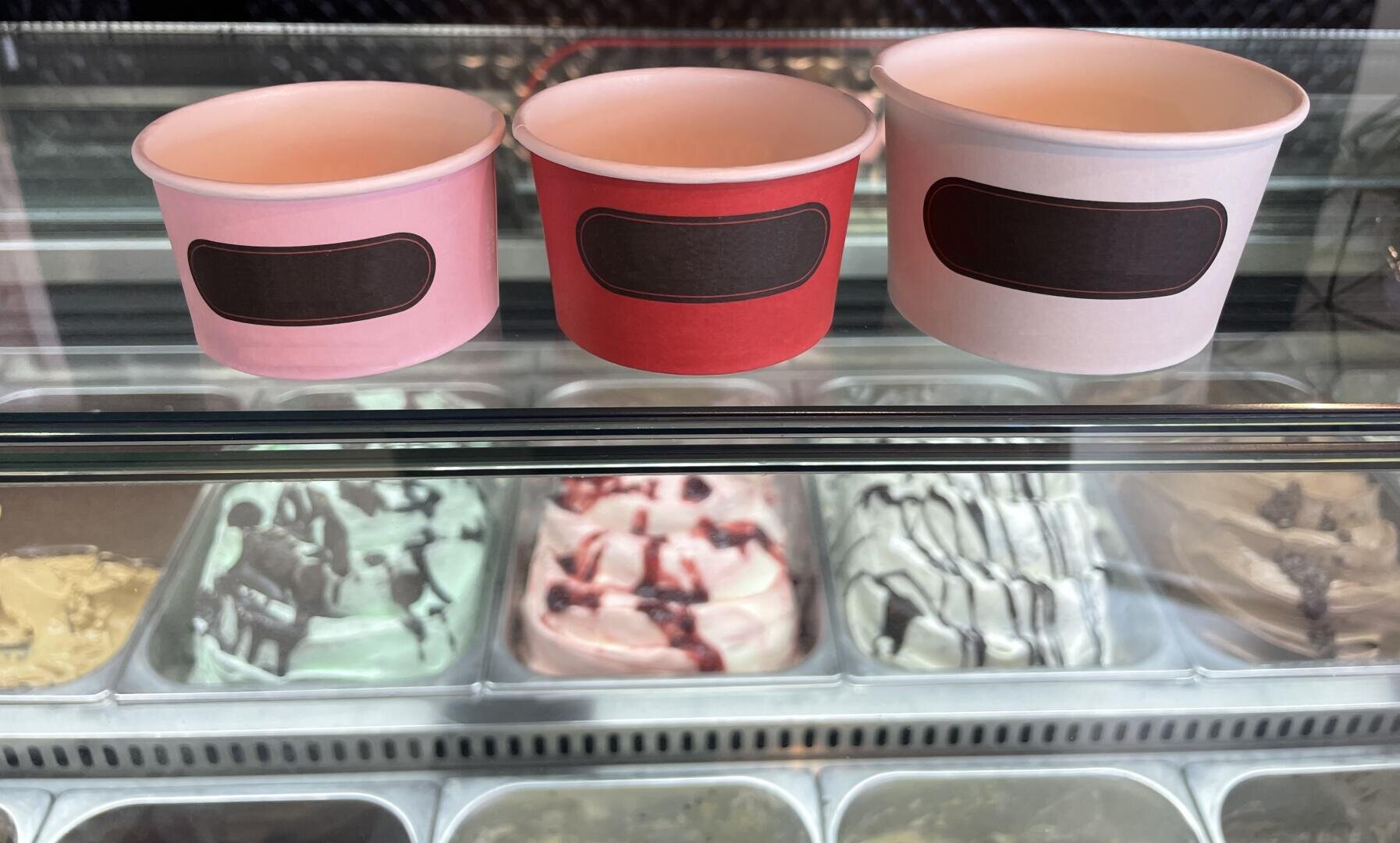 Three empty ice cream cups in pink, red, and white with blank black labels are displayed on a glass counter, with various flavors of ice cream in metal tubs visible underneath.