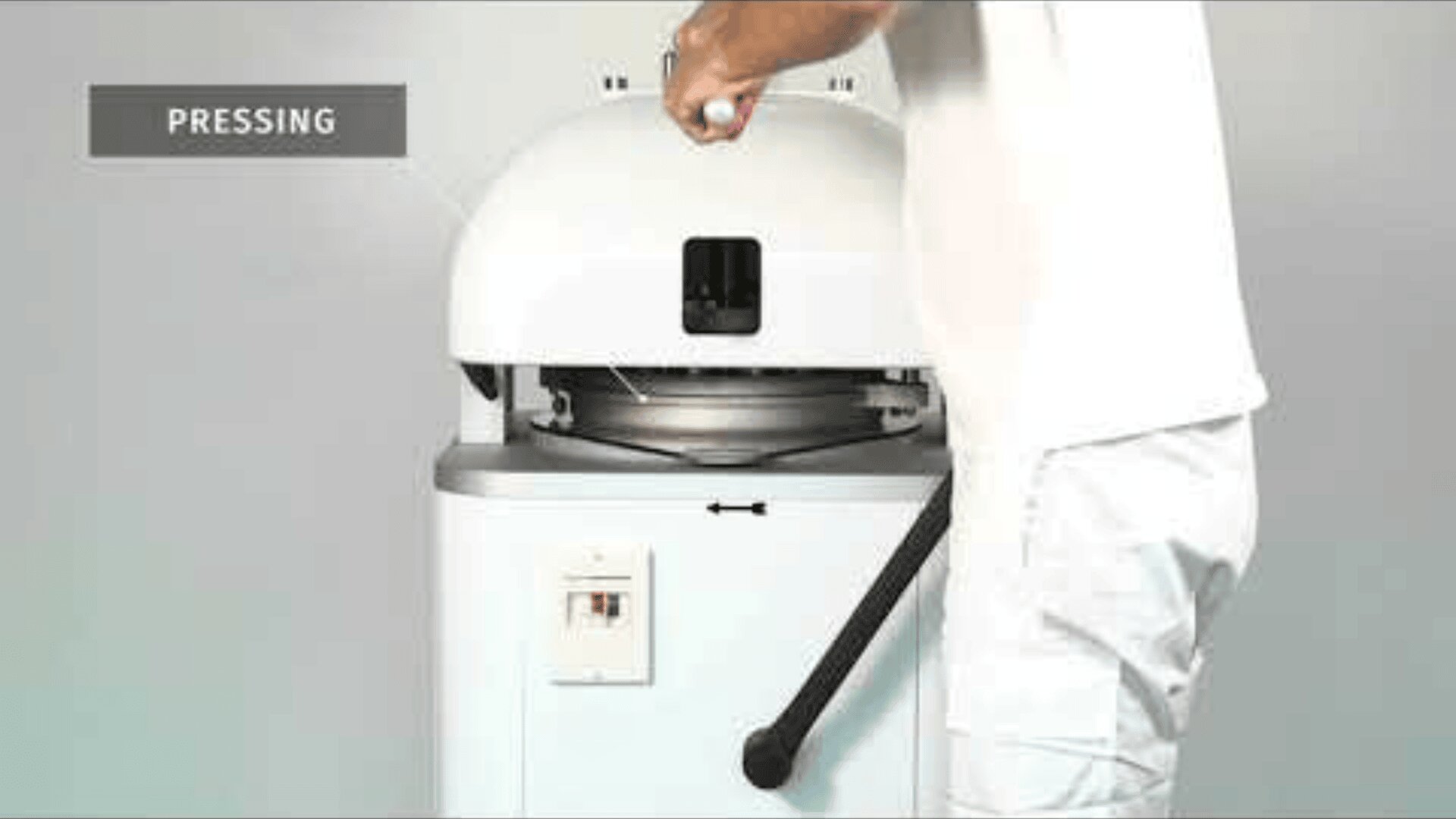 A person operates a white industrial machine by pressing a handle. The word PRESSING appears in a gray box in the upper left corner. The machine has a round, dome-shaped top and stands in front of a monochrome background.