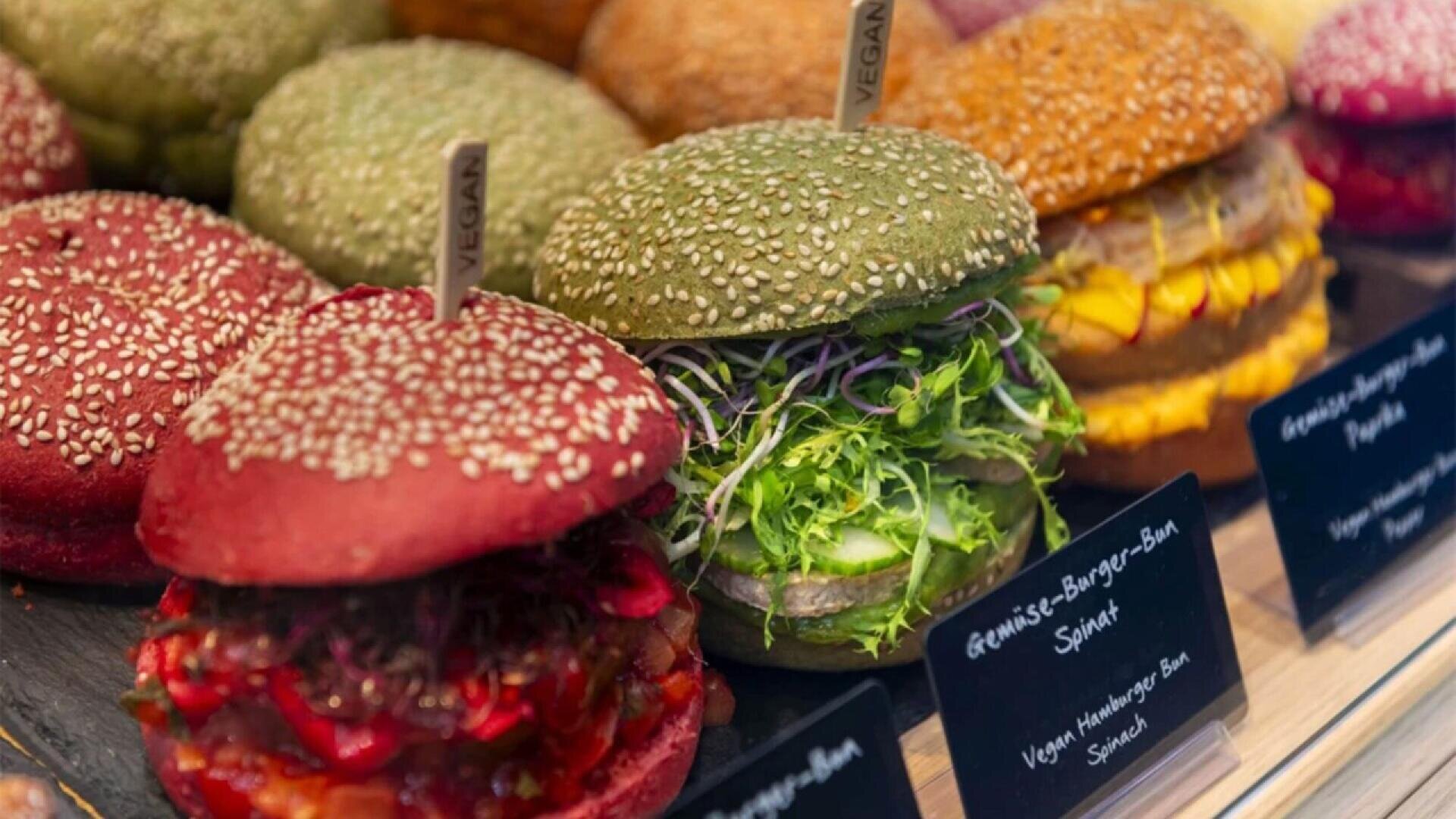 Colorful vegan burgers with red, green, and yellow seeded buns are displayed in a case, filled with fresh vegetables and greens. Small signs label each option, and wooden sticks marked vegan are inserted in the buns.