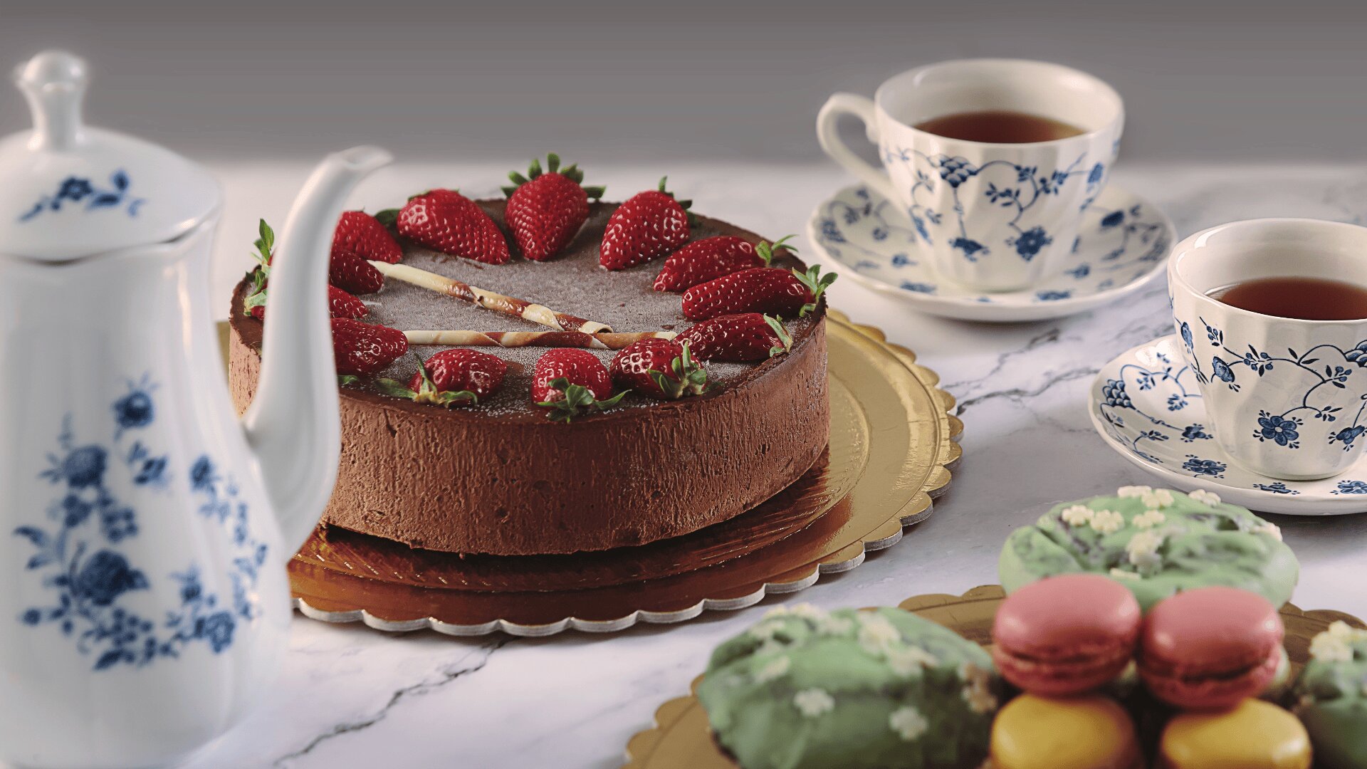 A chocolate cake topped with strawberries sits on a gold tray, surrounded by a teapot, two cups of tea in floral china, and plates of assorted green and pink macarons on a marble table.