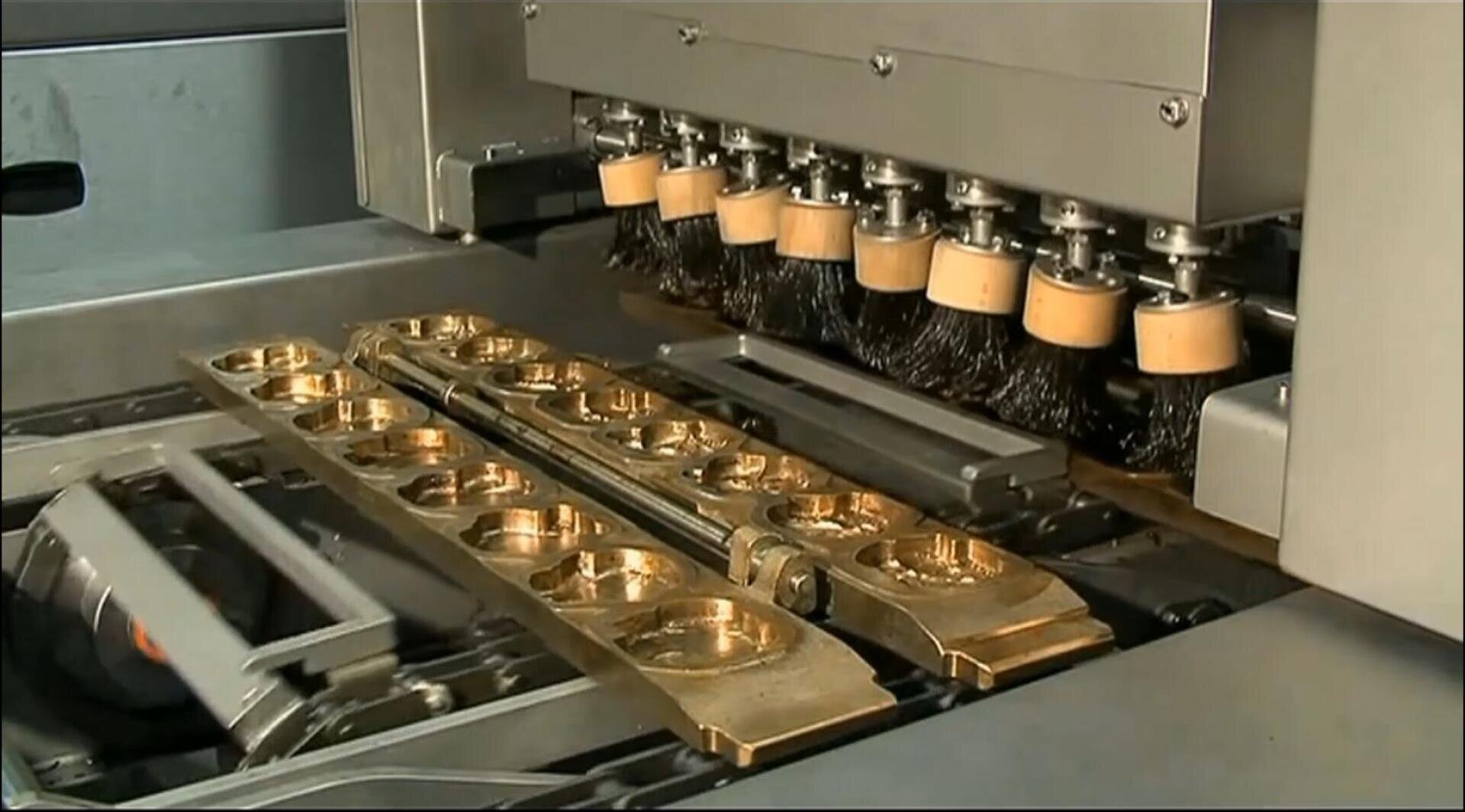 A machine with multiple rotating brushes polishes several gold-colored metal parts lined up in rows on a conveyor belt.