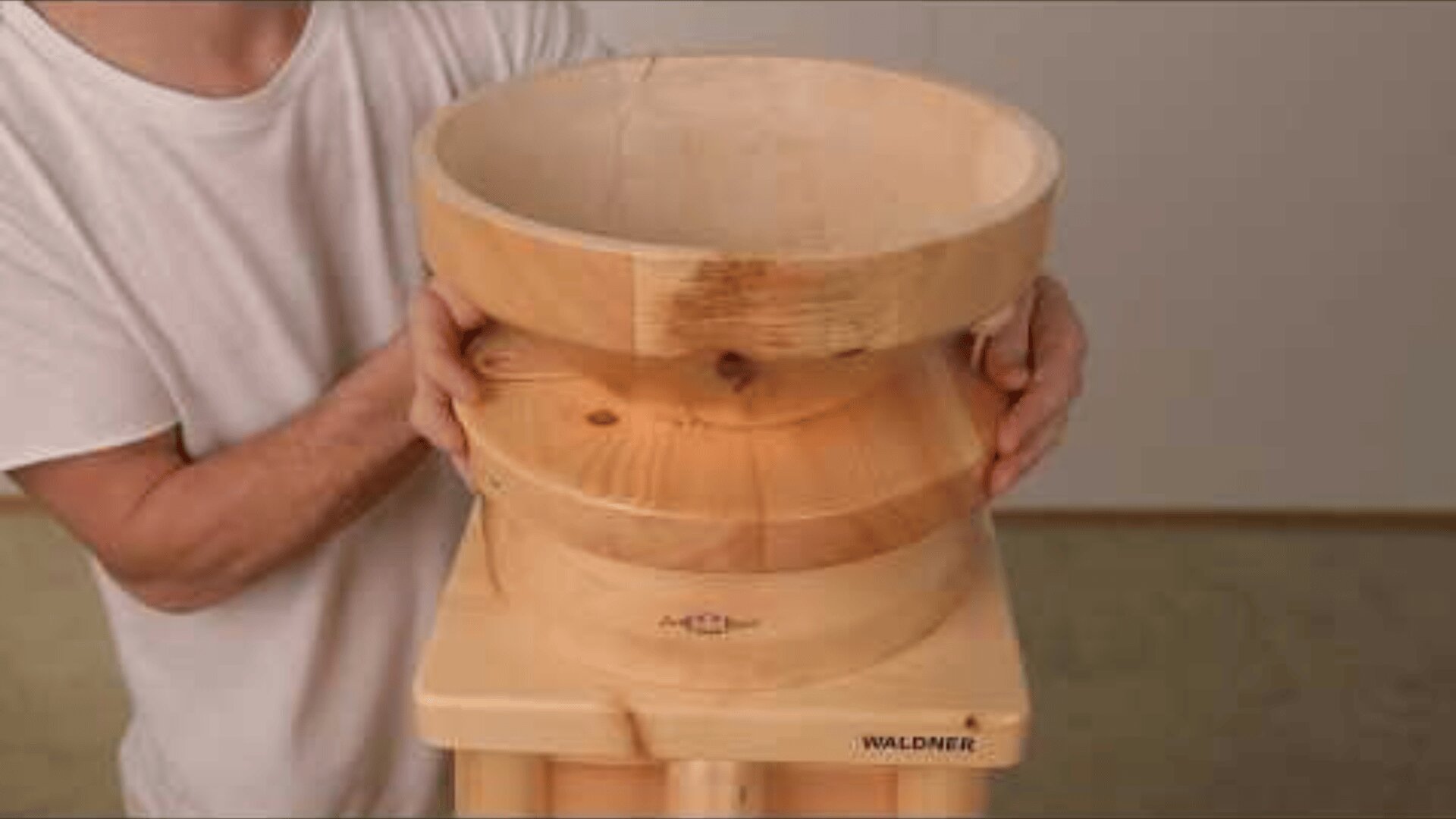 Eine Person in einem weißen Hemd baut eine Holzmühle mit der Gravur WALDNER auf dem Sockel zusammen oder hält sie in der Hand. Die Mühle hat eine glatte, natürliche Holzmaserung und steht auf einer hellen Fläche.