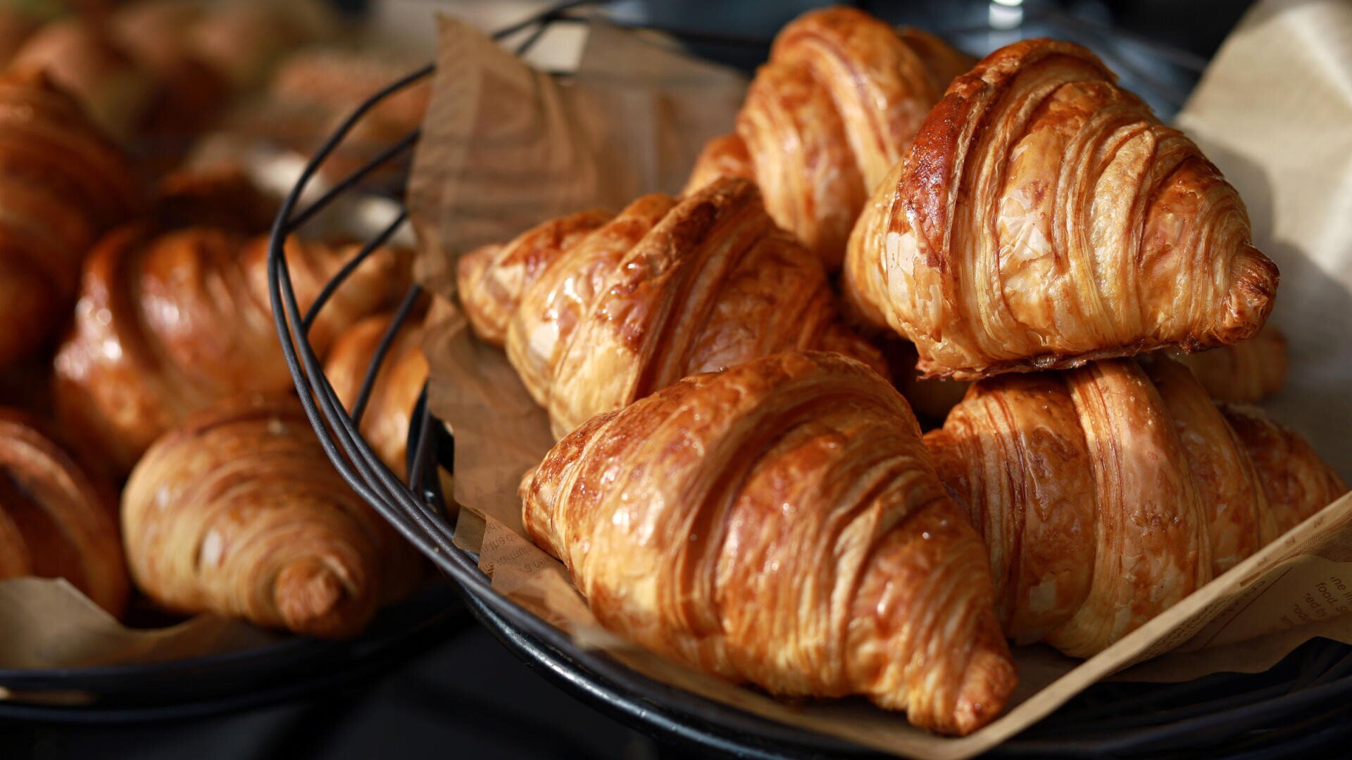 Ein Korb gefüllt mit goldbraunen, blättrigen Croissants auf braunem Pergamentpapier, im unscharfen Hintergrund sind weitere Gebäckstücke zu sehen.