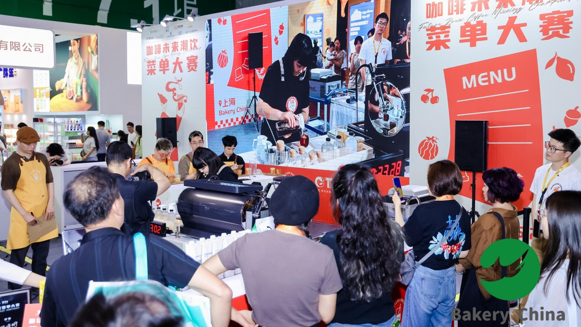 A crowd watches a baking demonstration at a trade show booth, with a chef preparing pastries while being filmed and shown on a large screen. The booth displays ingredients, utensils, and a bright red menu board.