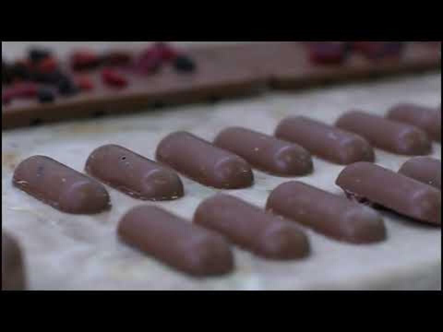 Rows of smooth, cylindrical milk chocolates are displayed on a light-colored surface, with more chocolates and small red pieces blurred in the background.