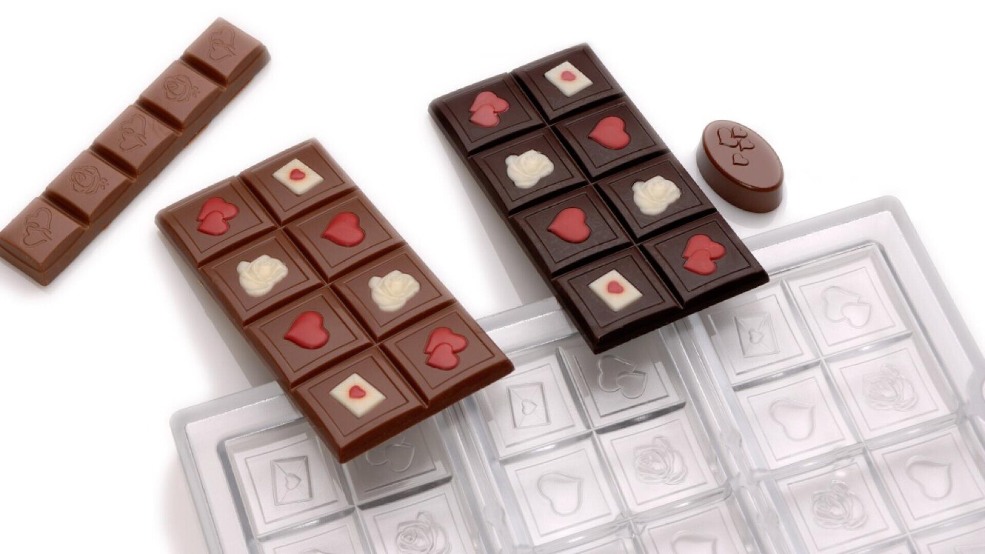 Milk and dark chocolate bars with heart and flower designs rest on a clear chocolate mold. One bar is segmented, with additional chocolate pieces nearby. The scene is set on a white background.