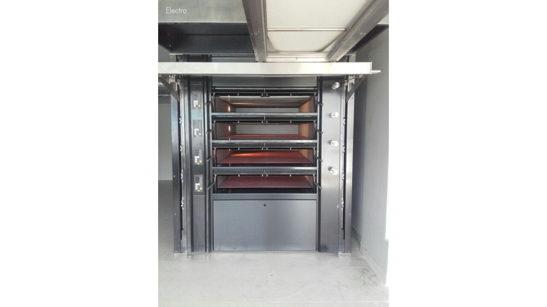A commercial bakery oven with four open baking chambers arranged vertically, each with a metal door and visible heating surfaces, set in a modern, clean kitchen space.
