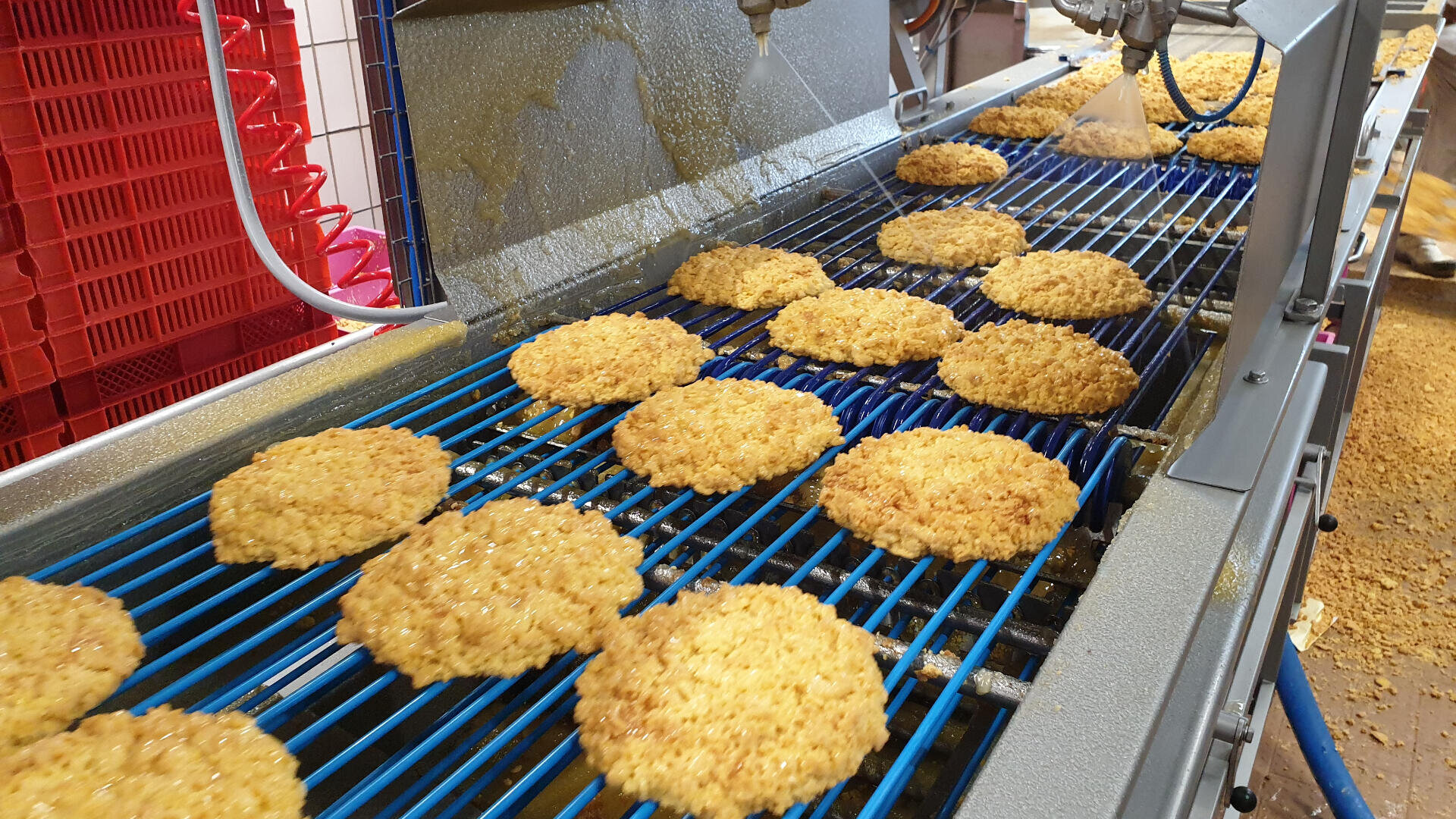 Breaded food patties move along a blue conveyor belt in a factory, with machines spraying liquid over them. Red crates are stacked in the background.