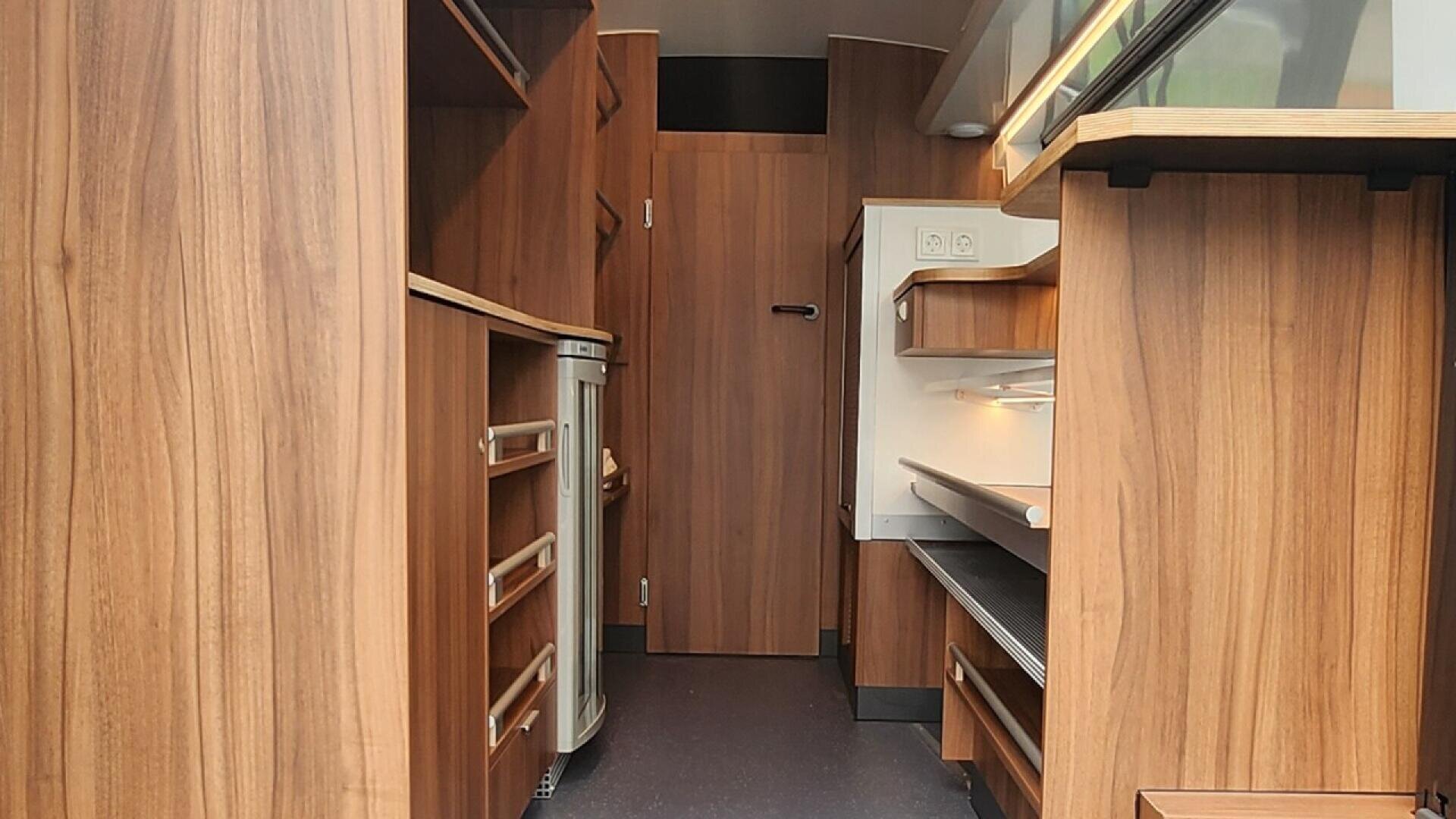 Interior of a compact, modern kitchen with wood-paneled cabinets, built-in shelves, drawers, and countertops. A small refrigerator and a closed door are visible at the end of the space.