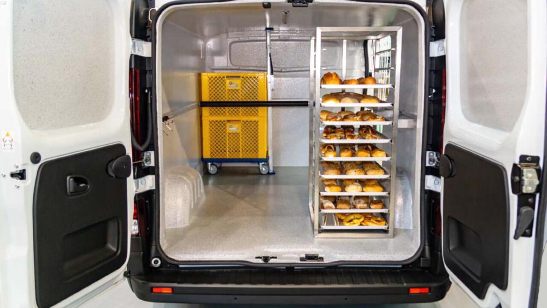 The open back doors of a refrigerated delivery van reveal racks of fresh bread and pastries on the right and a yellow rolling container at the back, all inside a clean, insulated storage compartment.