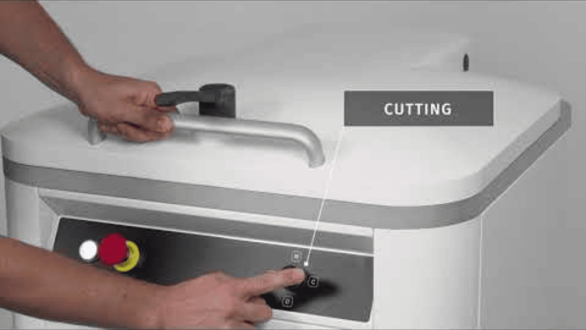 A person presses buttons on the control panel of a large white machine while holding its handle. A sign with the word CUTTING points to the control panel. The control panel has a yellow and red emergency stop button.
