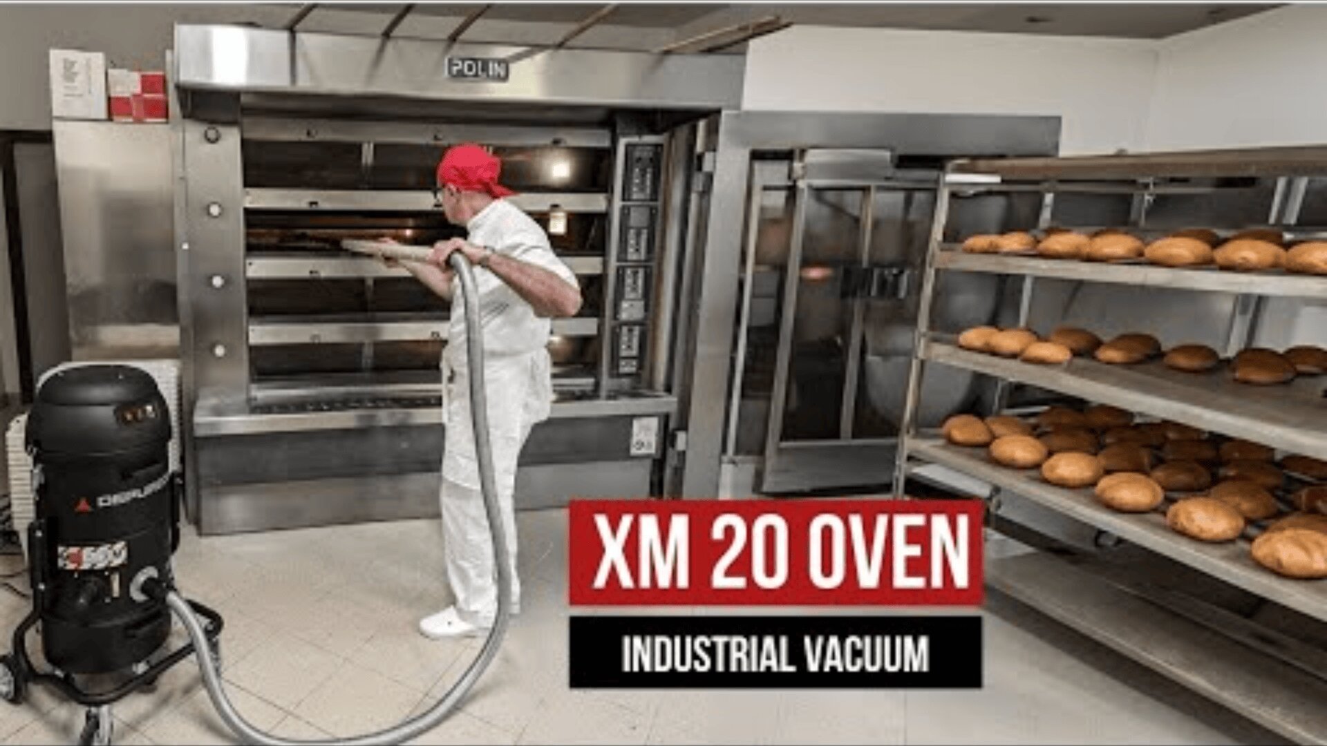 A baker in a white uniform and red cap uses a vacuum cleaner to clean a large industrial oven, near which there are shelves of freshly baked bread. The text reads XM 20 OVEN INDUSTRIAL VACUUM..