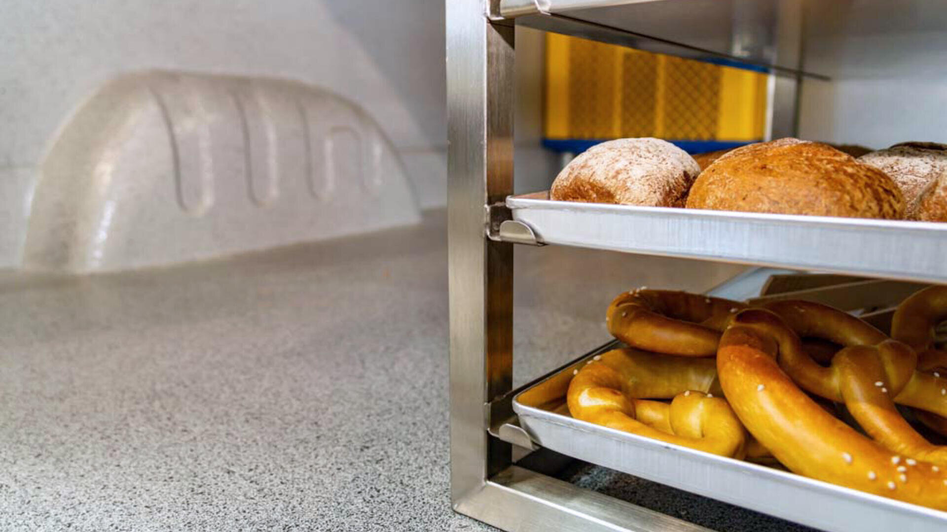 Ein Metallregal mit zwei Fächern hält runde Brötchen auf der oberen Ablage und weiche Brezeln auf der unteren Ablage, platziert auf einer gesprenkelten Arbeitsplatte in einer Küche oder Bäckerei.
