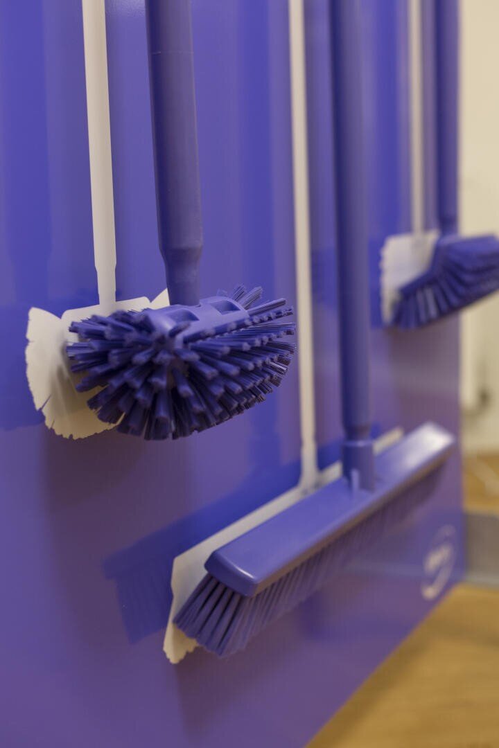 Close-up of blue cleaning brushes and brooms hanging neatly on a matching blue wall-mounted rack with white hooks.
