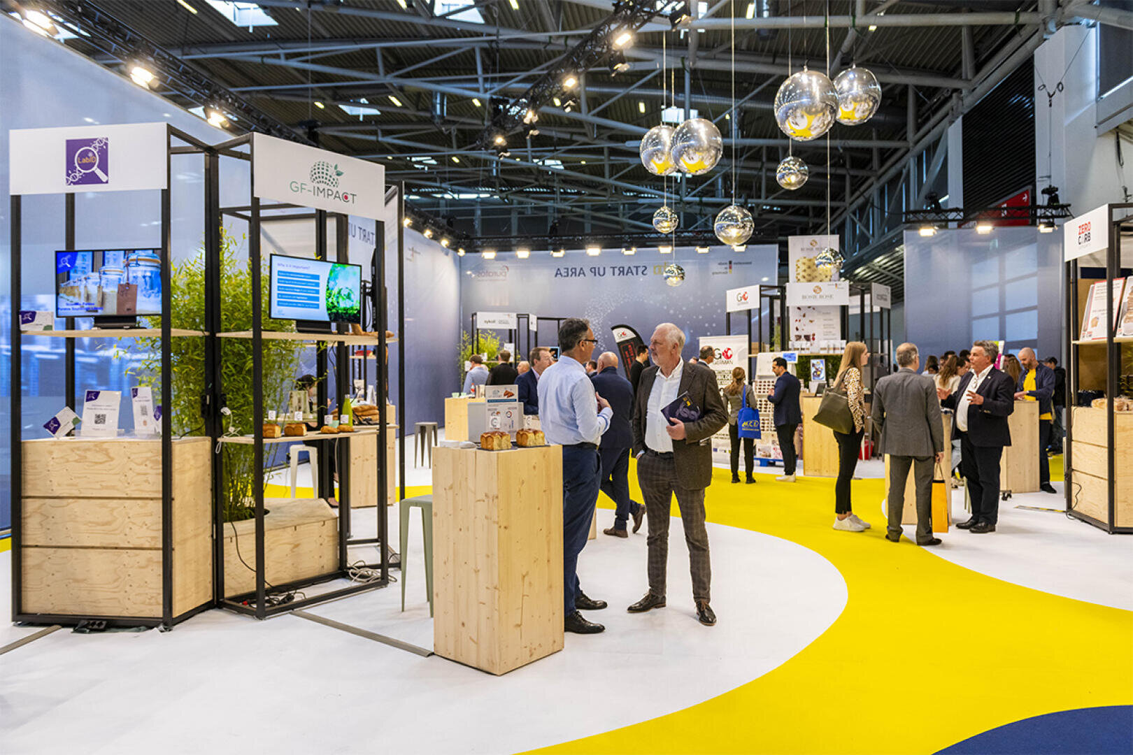 Auf einer modernen Indoor-Messe mit hölzernen Messeständen, digitalen Bildschirmen und hängenden Glasleuchten können sich Menschen austauschen und vernetzen. Der Boden ist in kräftigen Gelb-Weiß-Mustern gehalten, und an den Ständen sind Informationsmaterialien zu sehen.