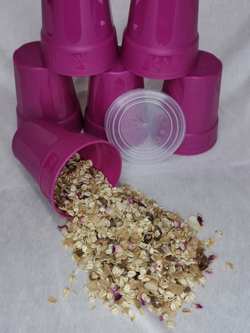 Sechs rosafarbene Plastikbecher sind gestapelt, wobei einer umgekippt ist und eine Mischung aus Haferflocken, Trockenfrüchten und Samen auf eine weiße Fläche verschüttet. Ein durchsichtiger Plastikdeckel liegt daneben.