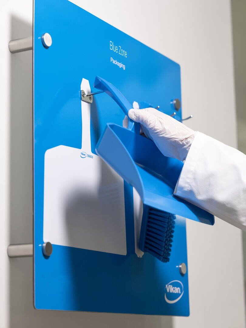 A gloved hand places a blue dustpan onto a blue wall-mounted cleaning station labeled Blue Zone Packaging, next to a white dustpan and blue brush.