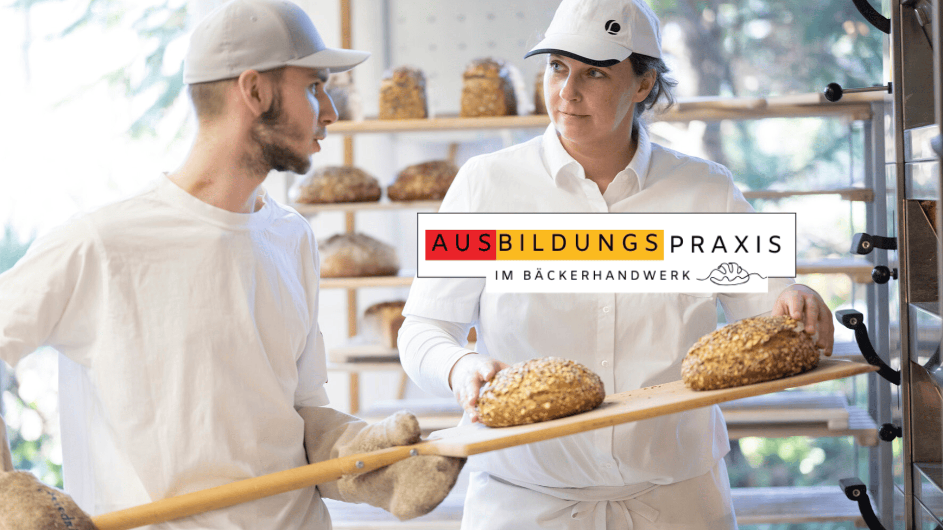 Zwei Bäcker in weißen Uniformen halten ein Holzbrett mit frischem Brot in einer Bäckerei. Im Hintergrund sind Regale mit Broten zu sehen. Der überlagerte Text lautet AUSBILDUNGSPRAXIS IM BÄCKERHANDWERK mit dem Logo eines Händedrucks.