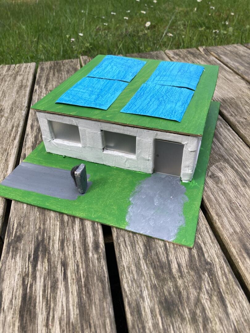 Ein kleines Modell eines Hauses mit grünen Wänden und blauen Solarmodulen auf dem Dach, auf einem grünen Sockel mit grauer Auffahrt und Briefkasten, platziert auf Holzbrettern im Freien mit Gras im Hintergrund.